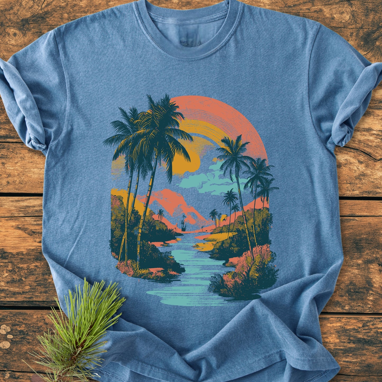 Along The River - Vintage Wash Tee features a retro tropical scene with palm trees, a lagoon, and mountains under a semicircular sunset, displayed on a heather-blue, soft-washed, ring-spun cotton T-shirt.