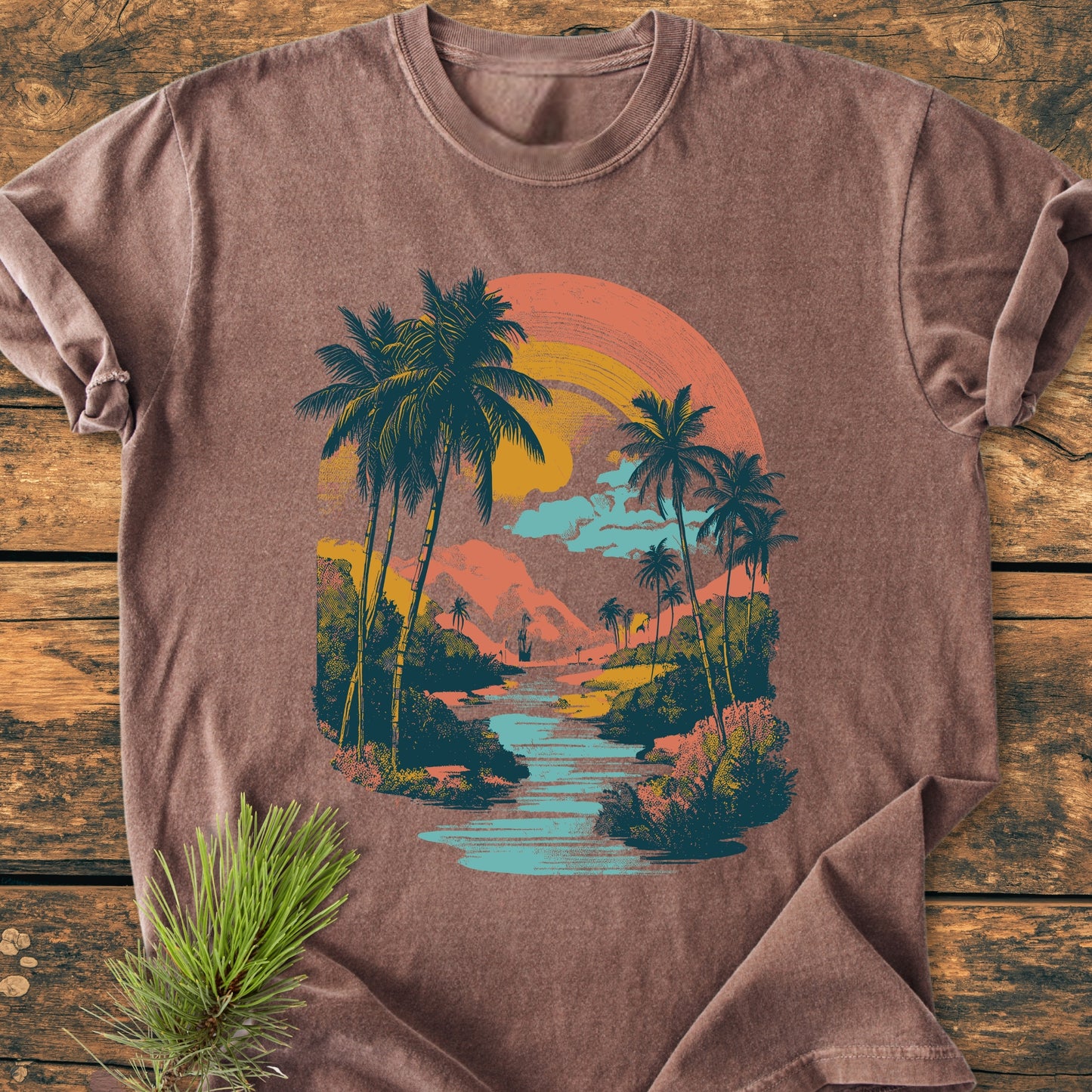 Along The River - Vintage Wash Tee displayed on wooden surface with a pine sprig, featuring a retro tropical sunset graphic with palm trees, a winding river, and a sun in warm tones.