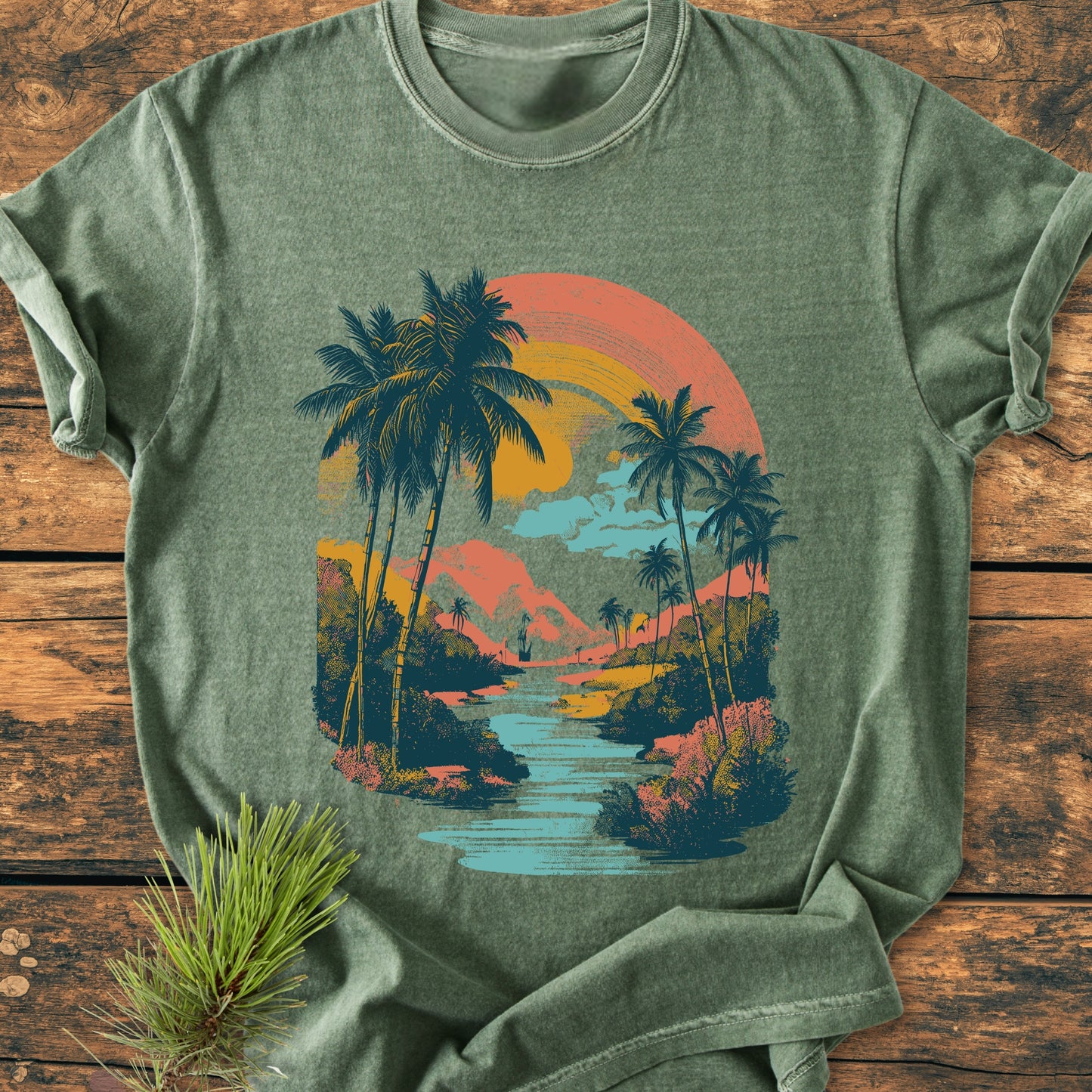 Along The River - Vintage Wash Tee, an olive-green T-shirt featuring a retro tropical scene with palm trees and a sunset, displayed flat on a wooden surface, highlighting its relaxed, soft-washed fabric.