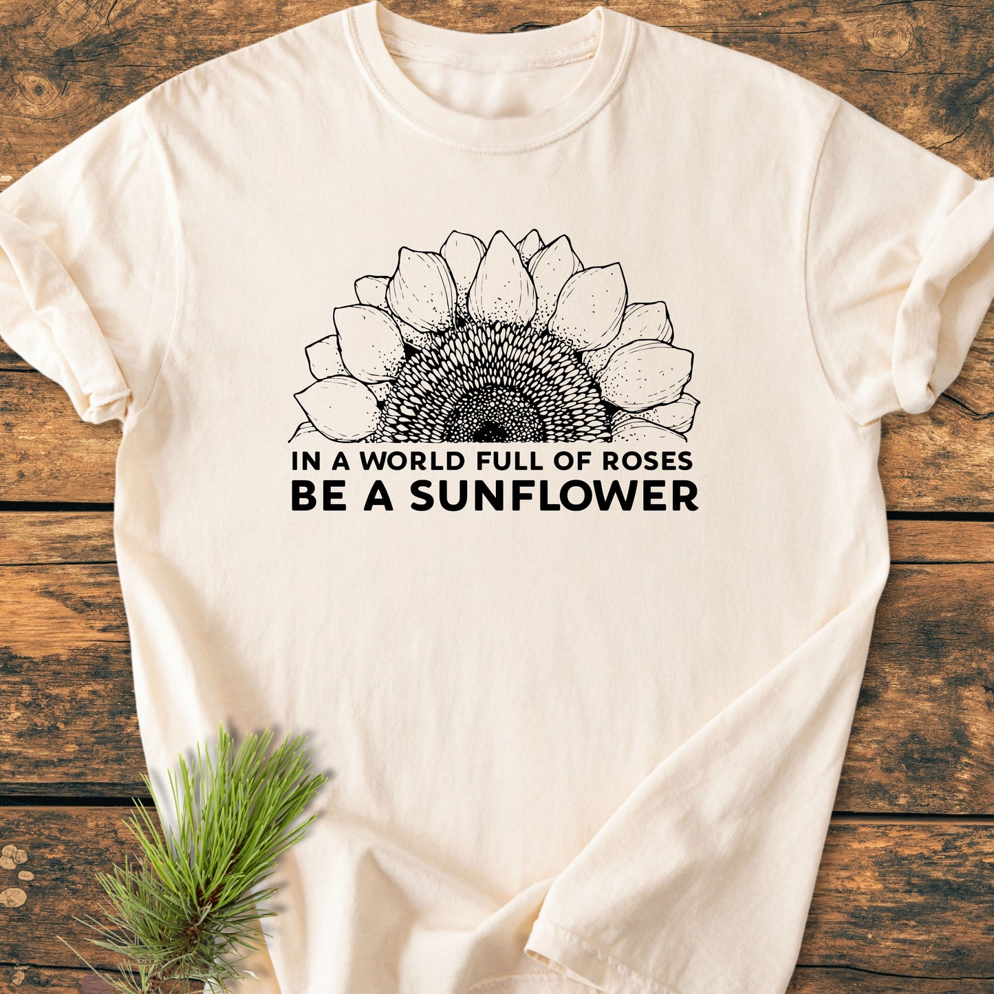 Be A Sunflower - Vintage Wash Tee features a half-sunflower line drawing with the slogan “In a world full of roses, Be a sunflower.” Laid on wood, accented by a pine sprig.
