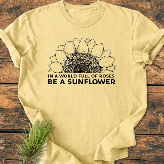 Be A Sunflower - Vintage Wash Tee features a half-sunflower graphic and slogan on a soft, relaxed-fit, pale yellow T-shirt, laid on wood, with pine needles accenting the scene.