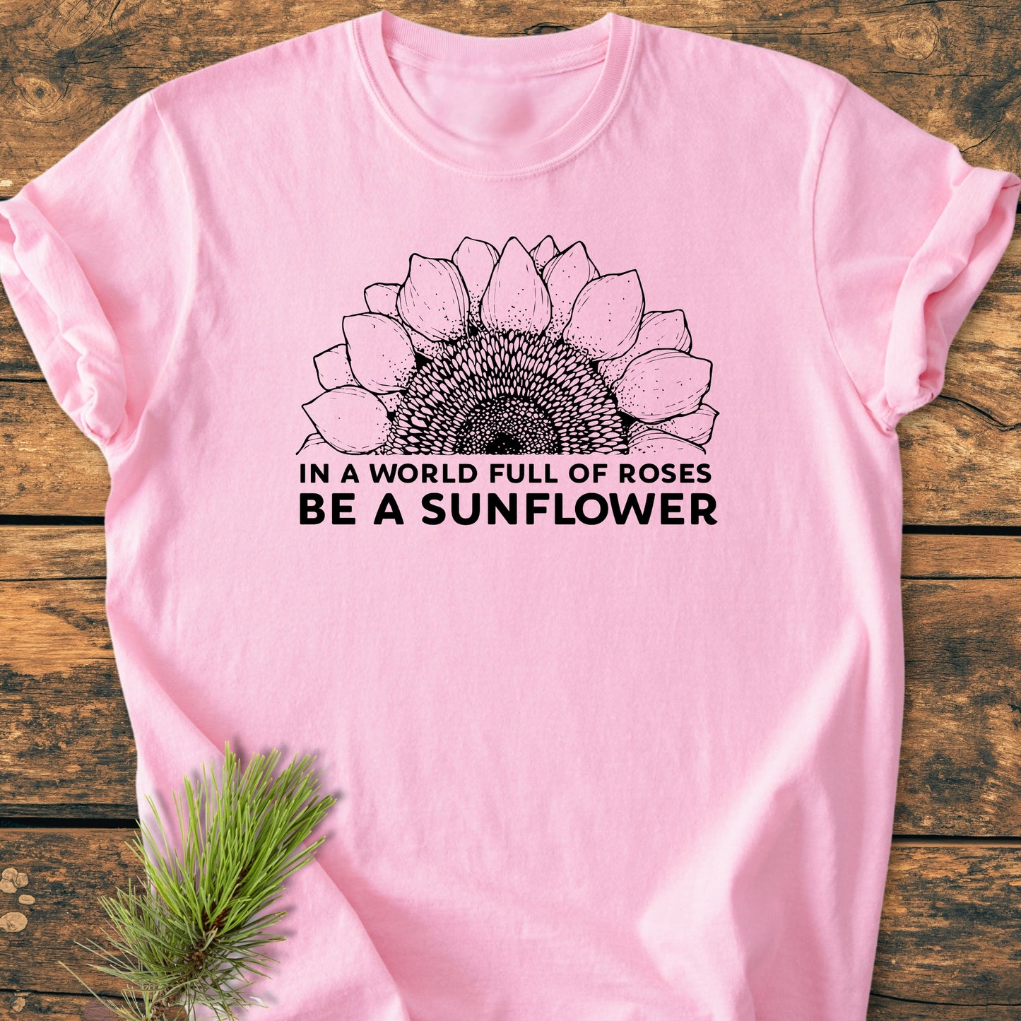 Be A Sunflower - Vintage Wash Tee features a sunflower illustration with a motivational slogan on a soft pink background, displayed on rustic wood with evergreen sprigs accentuating the design.