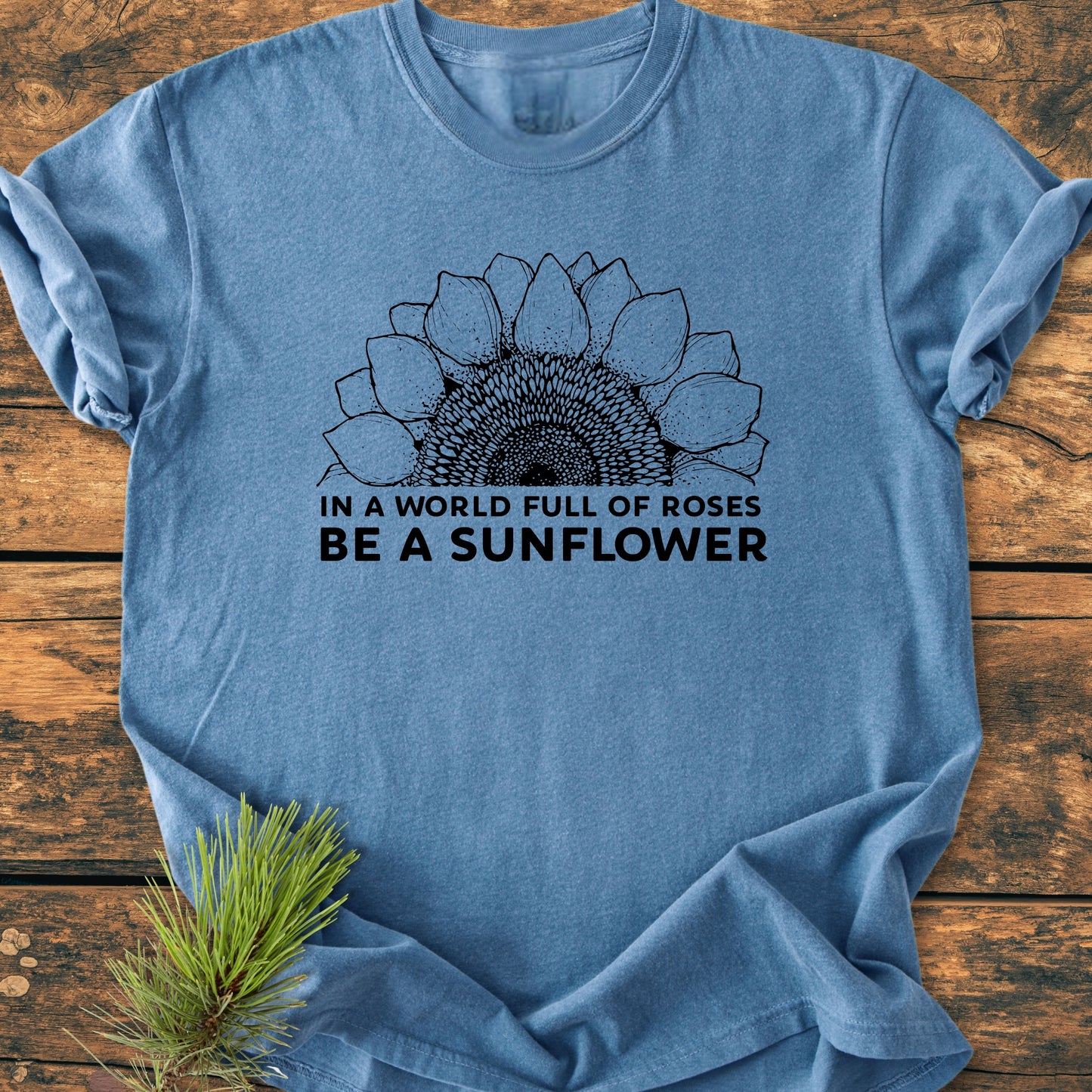 Be A Sunflower - Vintage Wash Tee features a sunflower illustration and motivational text on a light heather-blue shirt, laid flat on wood, emphasizing its cozy, relaxed fit and durable craftsmanship.