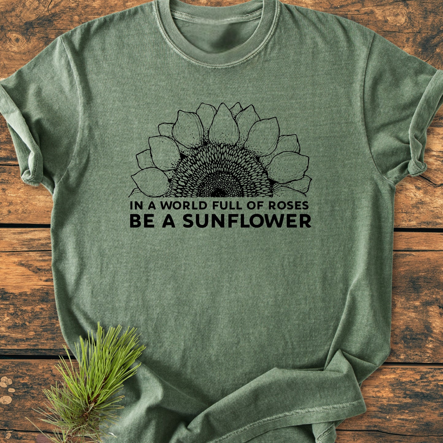 Olive-green Be A Sunflower - Vintage Wash Tee features a black sunflower line-drawing and slogan on soft, relaxed-fit fabric, laid on wood with a pine sprig accent.