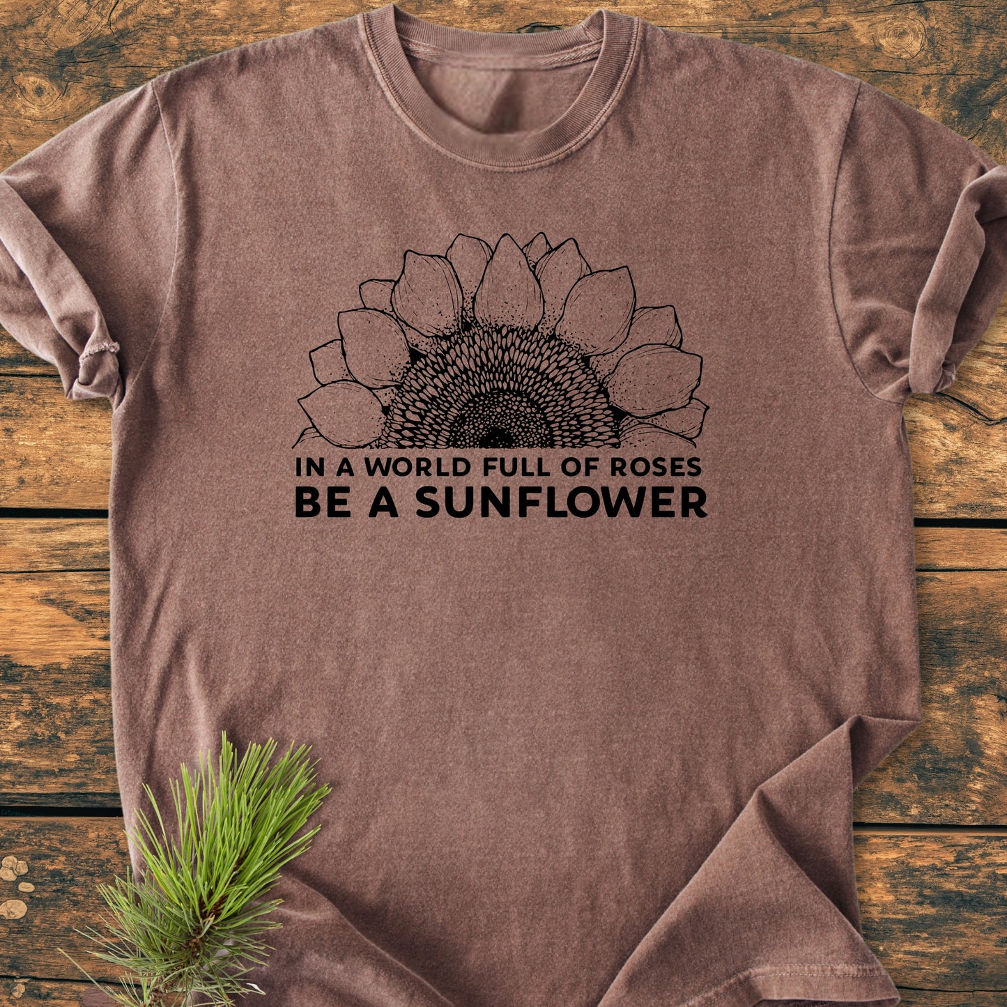Be A Sunflower - Vintage Wash Tee, featuring a half sunflower drawing and motivational quote, laid flat on a wooden surface with pine needles for a rustic touch.