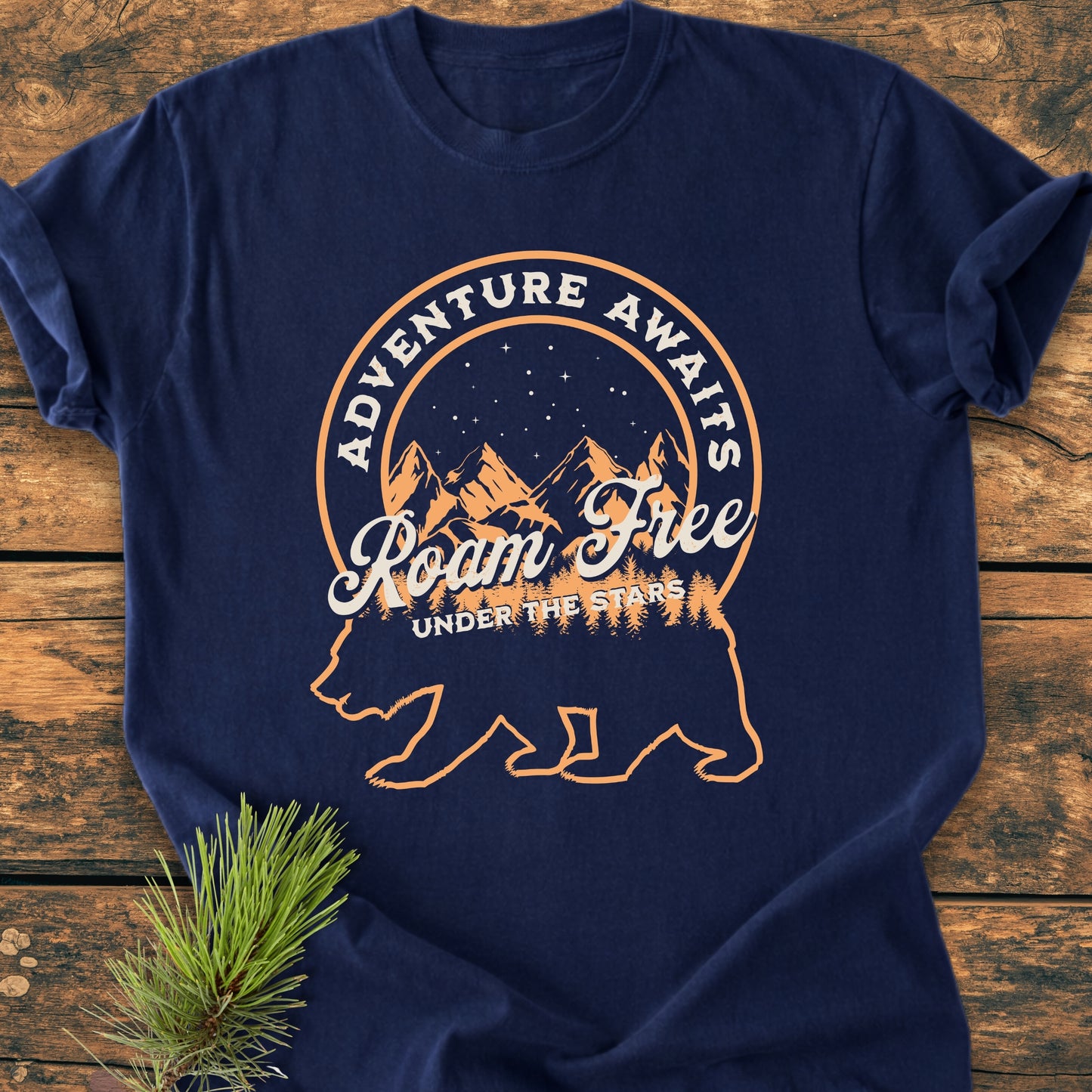 Roam Free Bear - Vintage Wash Tee featuring a navy-blue color with an orange-cream graphic of a bear, night sky, and slogans, laid out on a wooden surface with a pine sprig beside it.