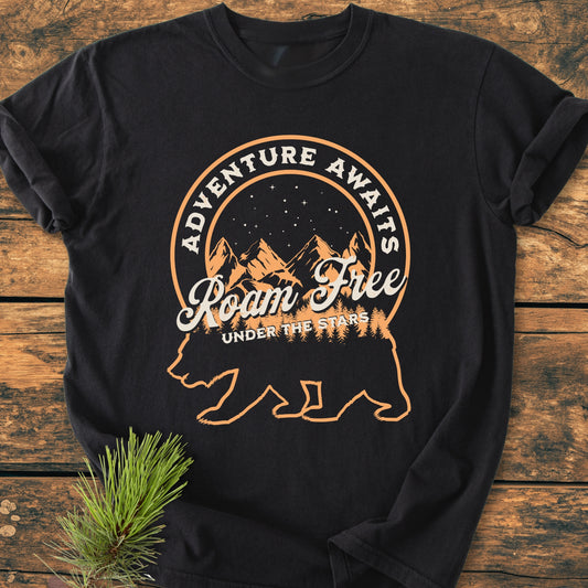 Roam Free Bear - Vintage Wash Tee features a black T-shirt with a bear silhouette containing a night-sky scene and adventure-themed text, laid on a wooden surface with a pine sprig.