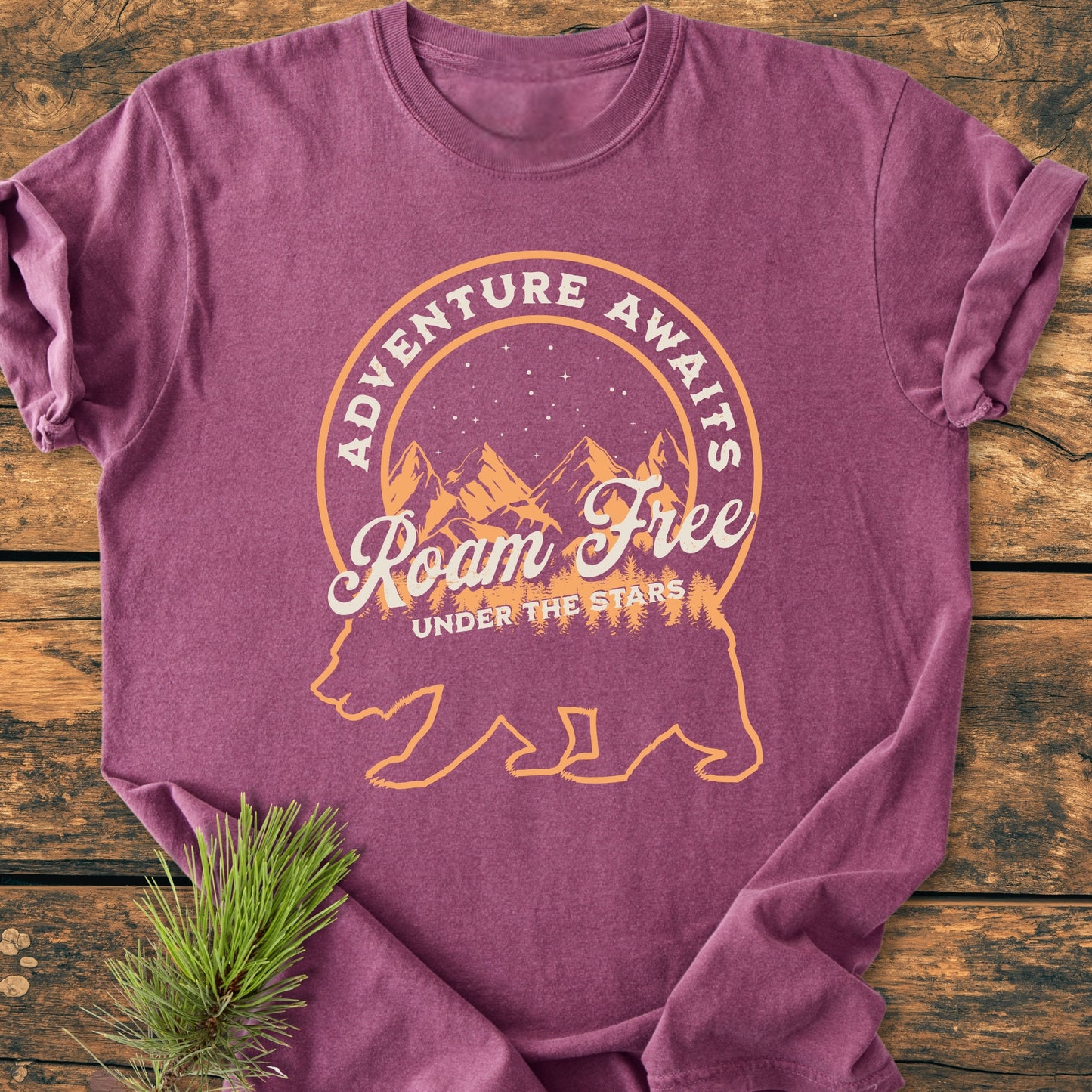 Roam Free Bear - Vintage Wash Tee features a bear silhouette with pine trees and mountains, adorned by ADVENTURE AWAITS. The heather-purple T-shirt lies folded on a wooden surface, exuding comfort and durability.
