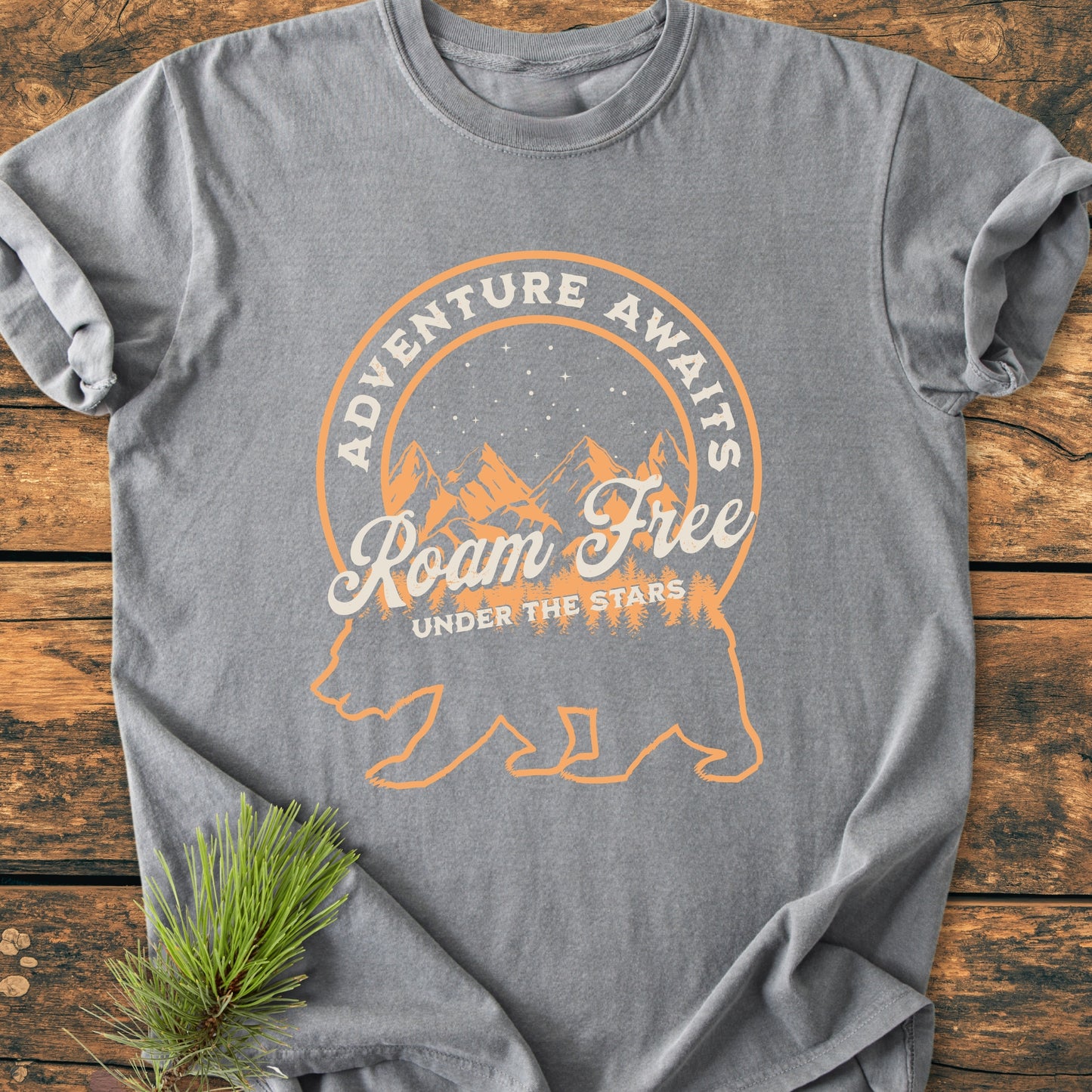 Roam Free Bear - Vintage Wash Tee: Gray T-shirt featuring a bear silhouette with a starry night, mountains, and trees design. Texts ADVENTURE AWAITS, Roam Free, and UNDER THE STARS accentuate this adventure-themed garment.