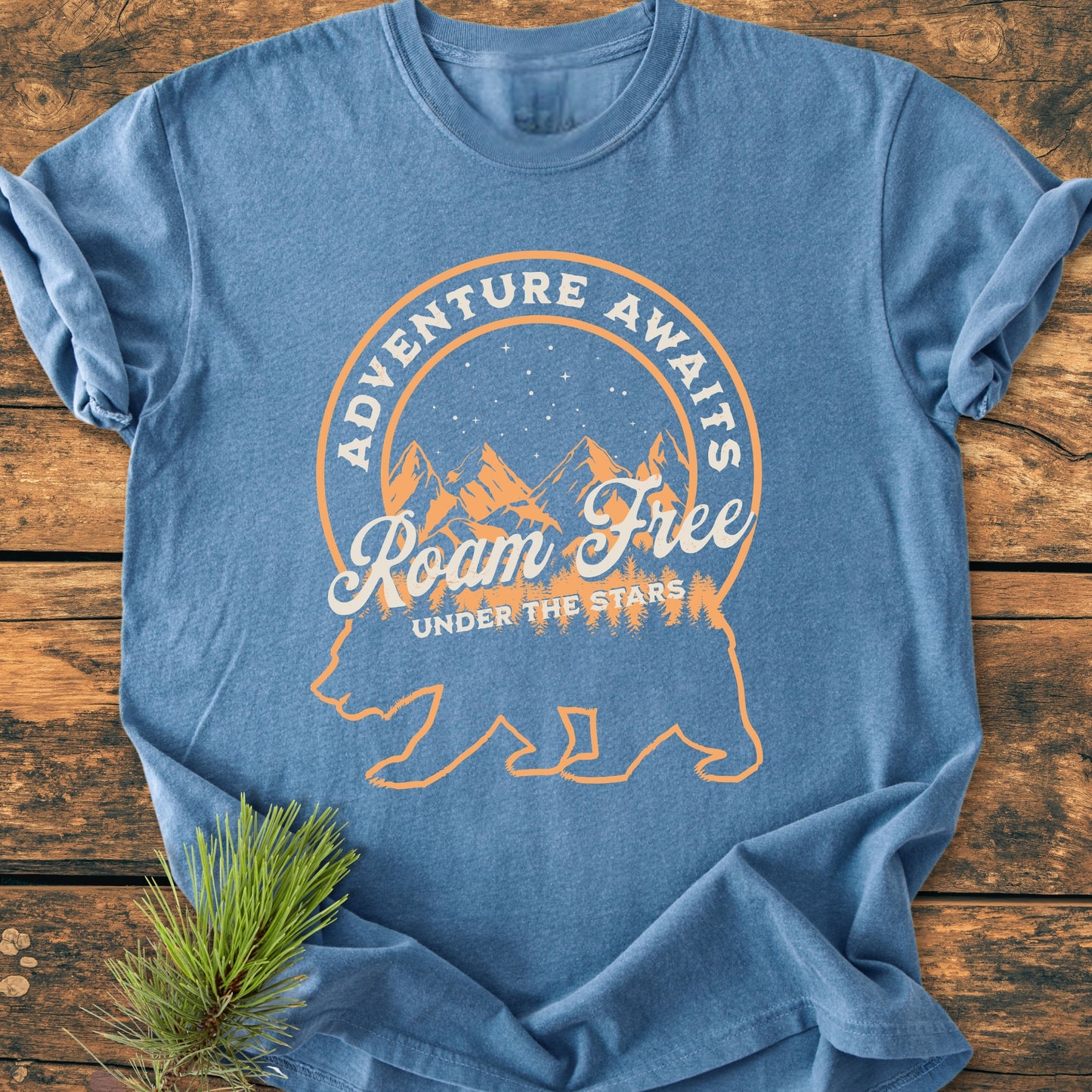 Roam Free Bear - Vintage Wash Tee: Heather-blue T-shirt featuring a graphic of a bear with pine trees, mountains, and a starry sky, accented by adventure-themed phrases. Laid flat with a pine sprig nearby.