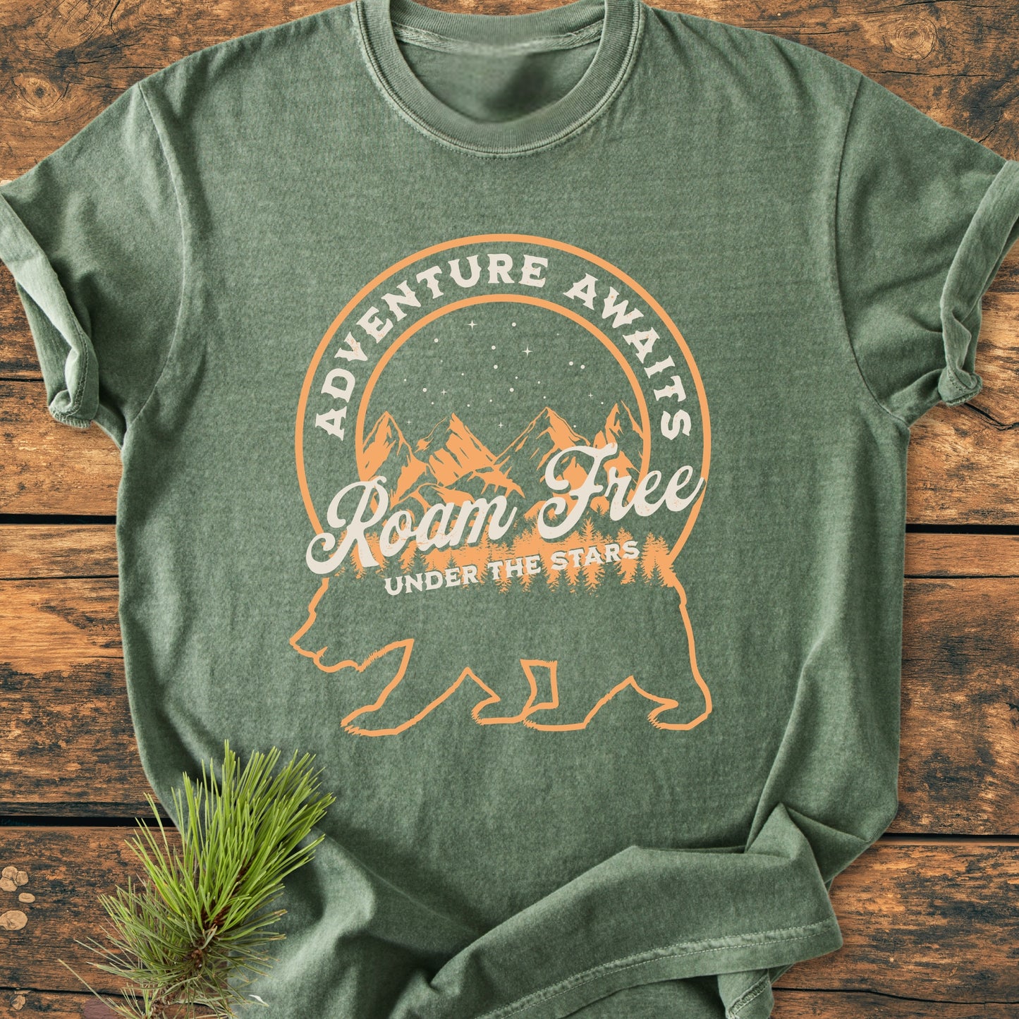 Roam Free Bear - Vintage Wash Tee: Heather-green T-shirt with a bear and mountain graphic, slogan “Adventure Awaits.” Laid flat with pine sprig accentuating its rustic, nature-inspired design.