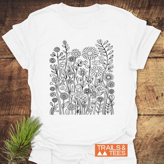 Wildflower Trail T-Shirt featuring a black line-drawing of wildflowers on the front, laid on a wood surface with a small TRAILS & TEES logo in the corner, and a sprig of pine needles beside it.