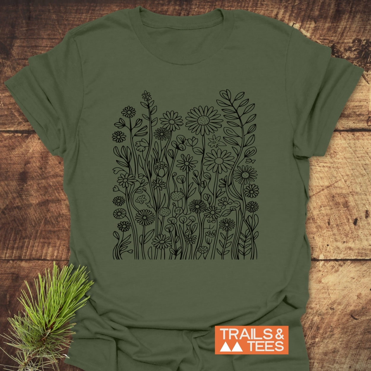 Wildflower Trail T-Shirt featuring a black line-art garden of daisies and leafy sprigs on an olive-green fabric, showcasing a unisex fit ideal for adventures. Photographed on a wooden surface with a “TRAILS & TEES” logo.