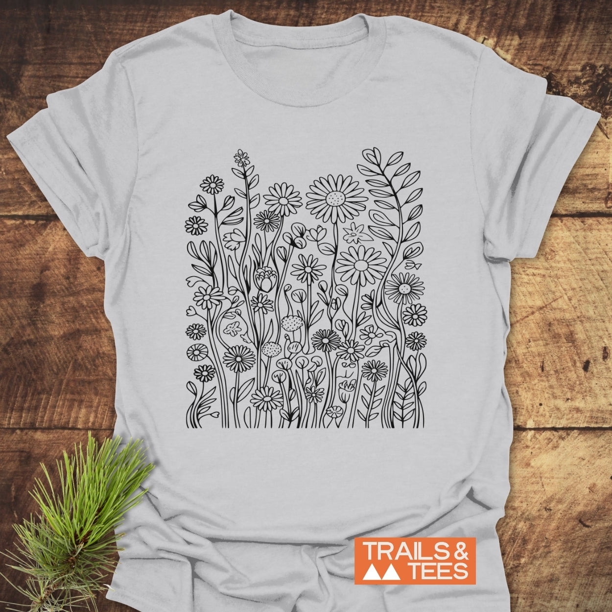 Wildflower Trail T-Shirt features a black line-drawing of wildflowers on white cotton, laid on wood. TRAILS & TEES text is visible with a pine sprig beside it. Designed for comfort and adventure.