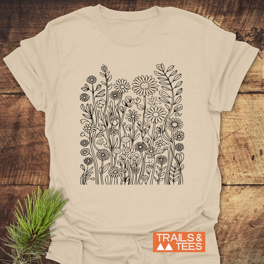 Wildflower Trail T-Shirt flat-lay showcasing black line-drawing of wildflowers on light-beige fabric. Features a small “TRAILS & TEES” logo and a pine branch accent on a wooden surface.