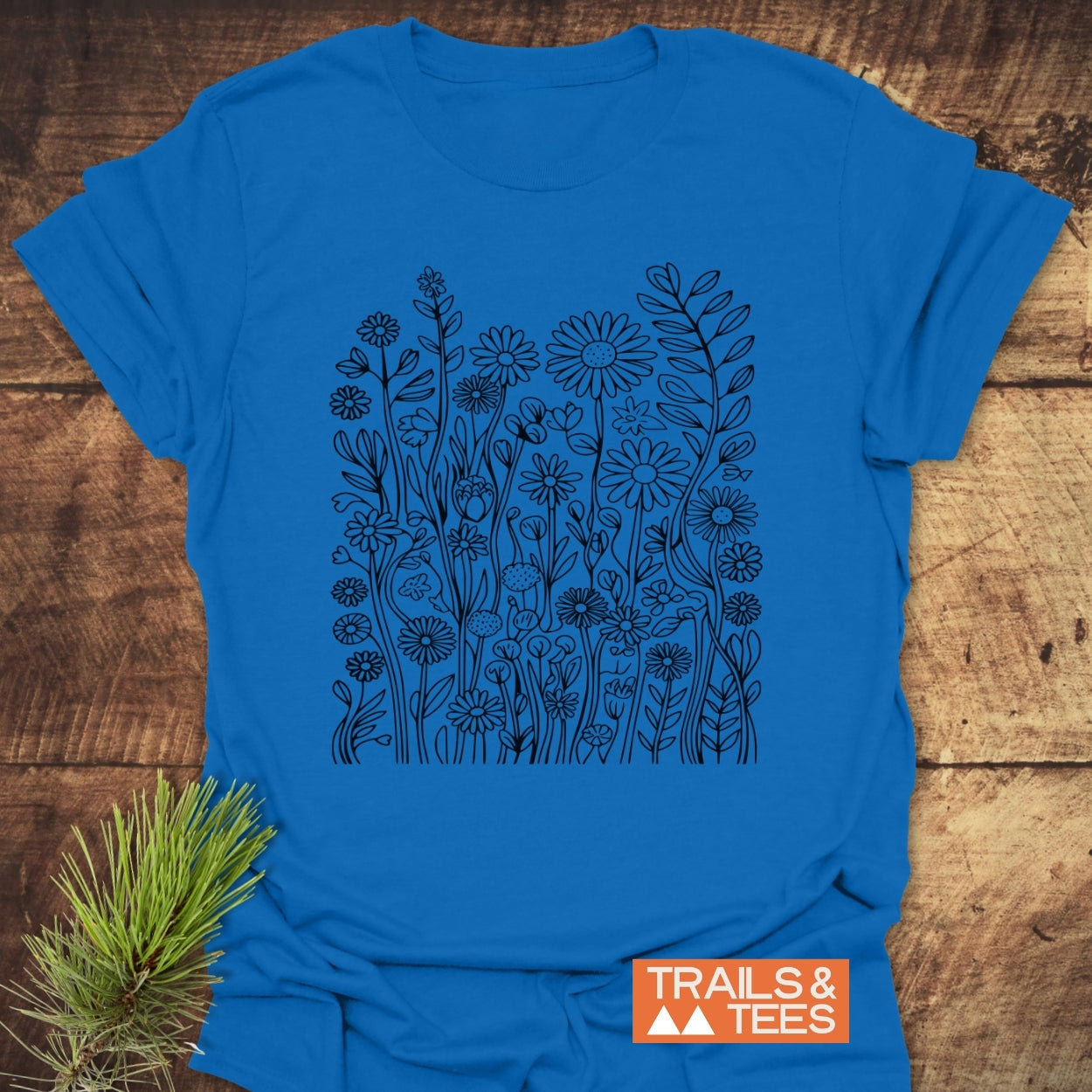 Wildflower Trail T-Shirt showcasing a black line-drawing of meadow blooms on a bright blue fabric, designed for comfort and adventure. Features a TRAILS & TEES logo and a pine twig accent.