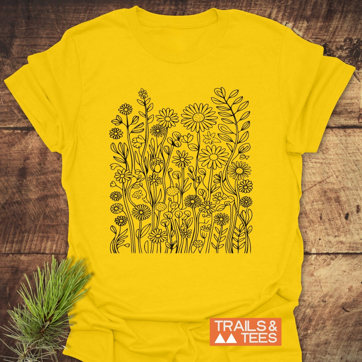 Wildflower Trail T-Shirt featuring a black, hand-drawn wildflower illustration on the front. Set against a wooden surface, the unisex tee is designed for comfort and adventure, featuring the TRAILS & TEES logo.