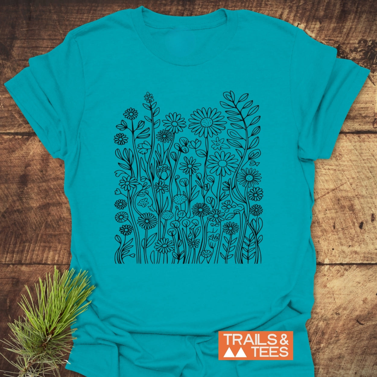 Wildflower Trail T-Shirt featuring black line-art wildflowers and leaves design, orange TRAILS & TEES label, on wooden background with pine sprig, ideal for adventure with soft, lightweight cotton.