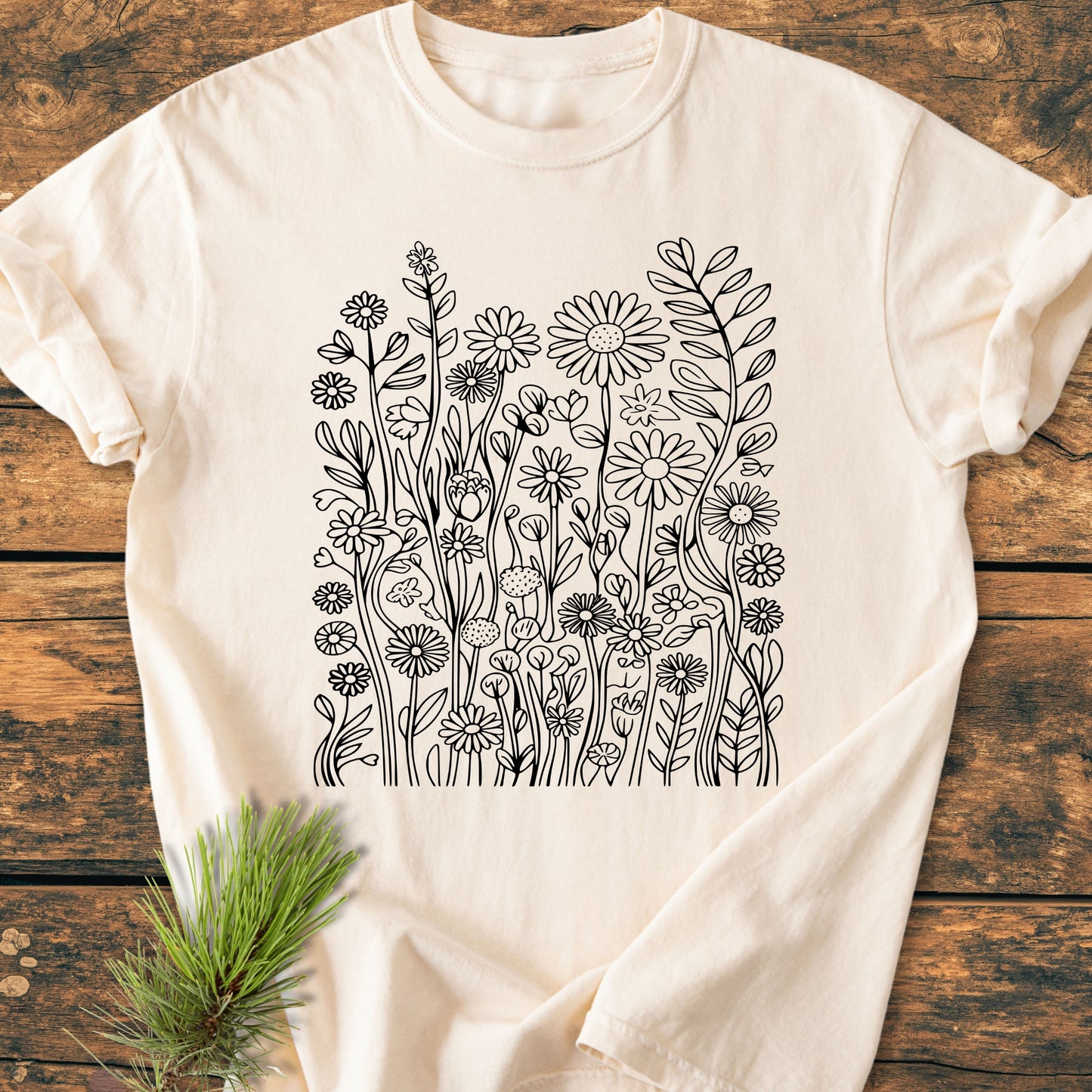 Wildflower Trails - Vintage Wash Tee featuring a black line-art of wildflowers on a light-cream T-shirt, laid on a rustic wooden surface with a pine sprig nearby.