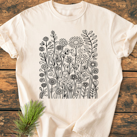 Wildflower Trails - Vintage Wash Tee featuring a black line-art of wildflowers on a light-cream T-shirt, laid on a rustic wooden surface with a pine sprig nearby.