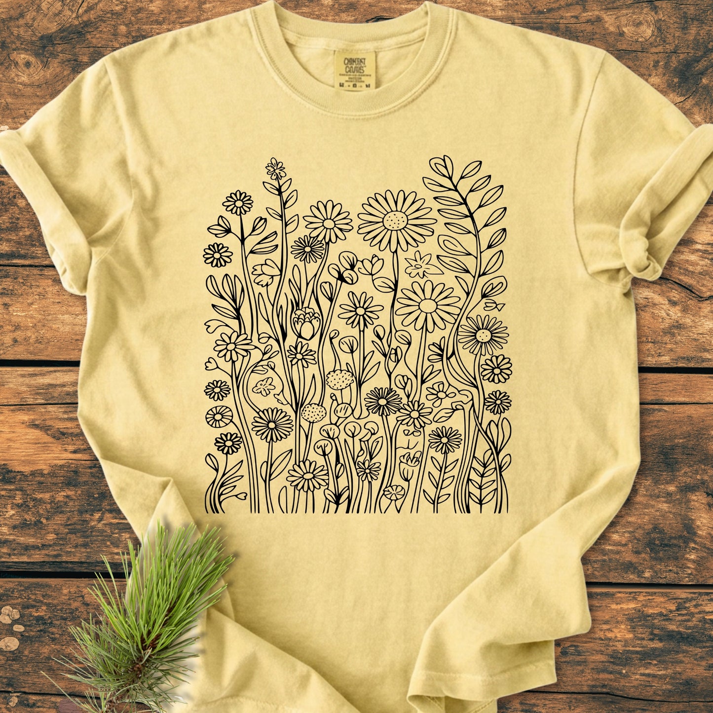 Wildflower Trails - Vintage Wash Tee, featuring intricate black wildflower line-art on a light yellow fabric. Displayed on a wooden surface with a pine sprig accent, highlighting its relaxed and durable design.