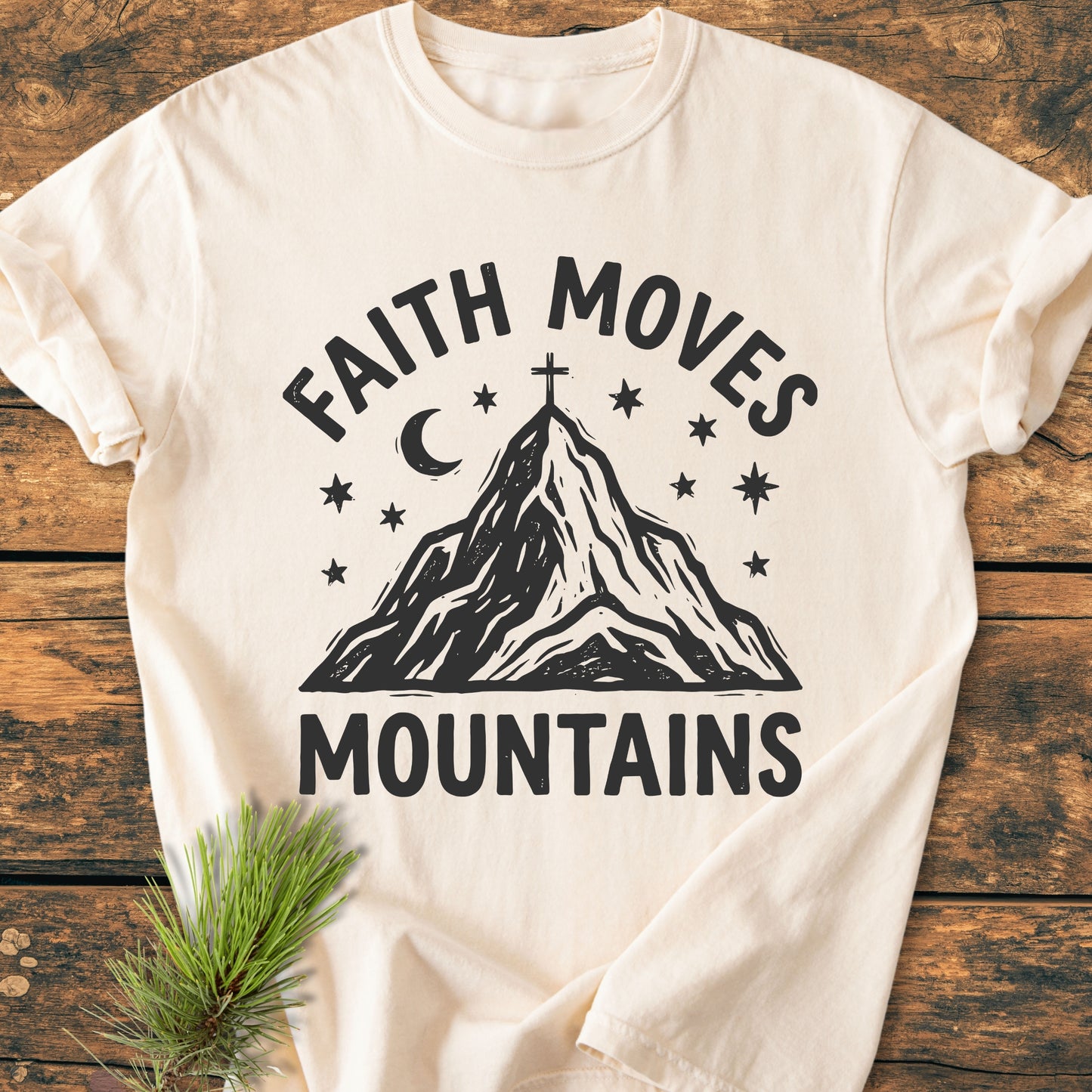 Faith Moves Mountains - Vintage Wash Tee featuring a mountain graphic with stars and crescent moon, highlighted by FAITH MOVES and MOUNTAINS text; laid on wood, accompanied by a pine sprig.