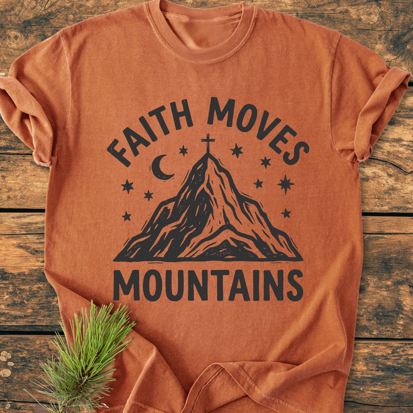 Faith Moves Mountains - Vintage Wash Tee shown on a wooden surface, featuring a mountain peak design with a cross, moon, and stars. Rolled sleeves and a pine sprig enhance its rustic appeal.