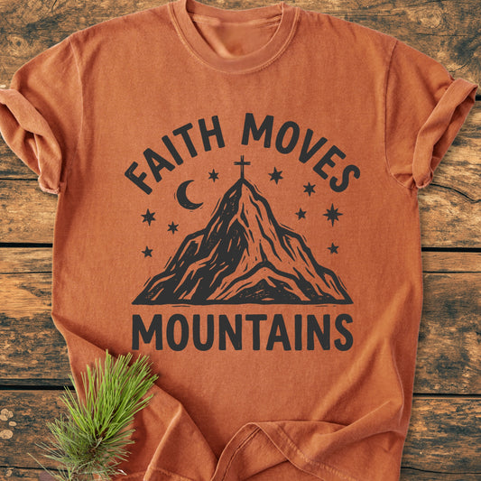 Faith Moves Mountains - Vintage Wash Tee shown on a wooden surface, featuring a mountain peak design with a cross, moon, and stars. Rolled sleeves and a pine sprig enhance its rustic appeal.