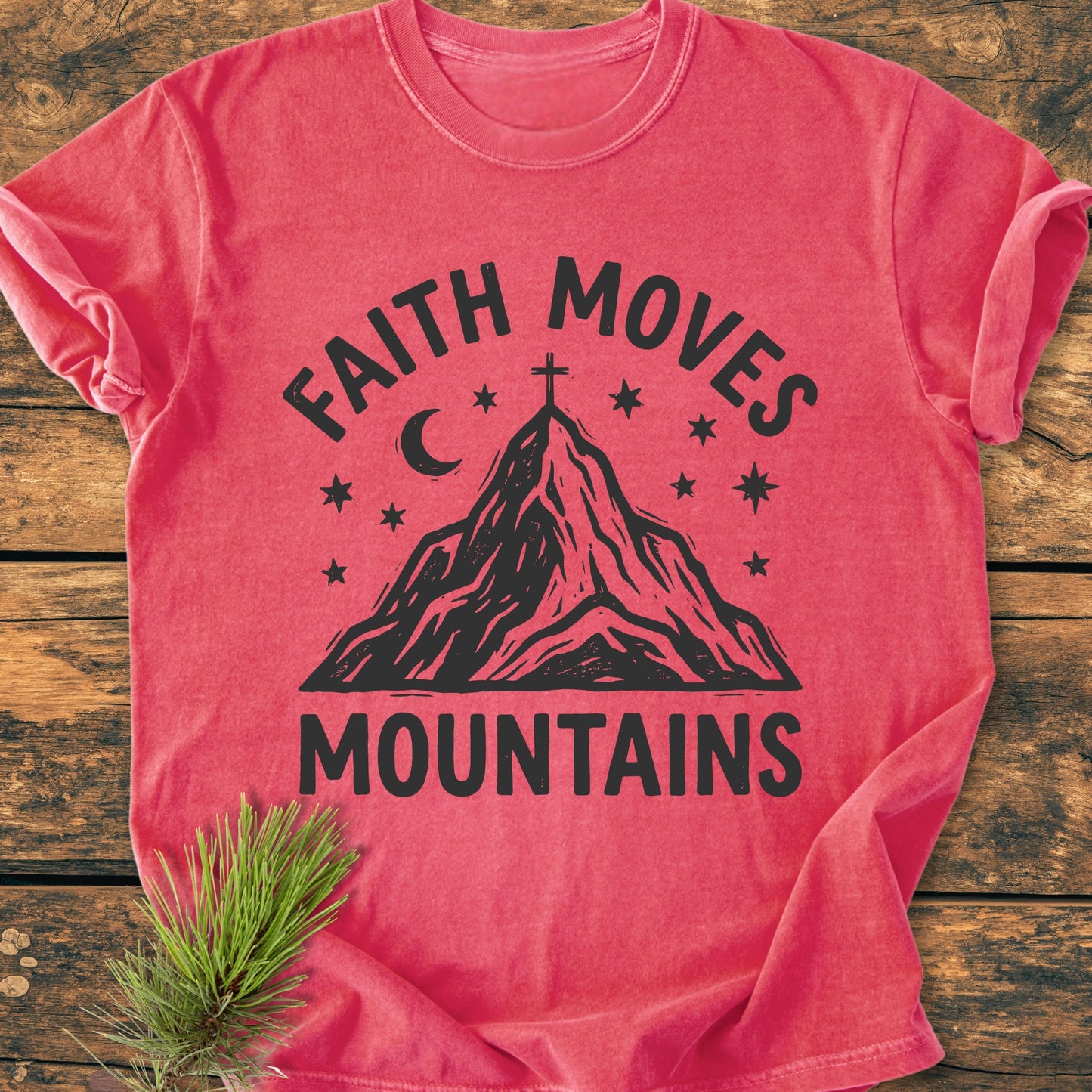 Faith Moves Mountains - Vintage Wash Tee features a stylized mountain with a cross, stars, and crescent moon, printed on a red T-shirt, laid on a wooden surface with a pine sprig.
