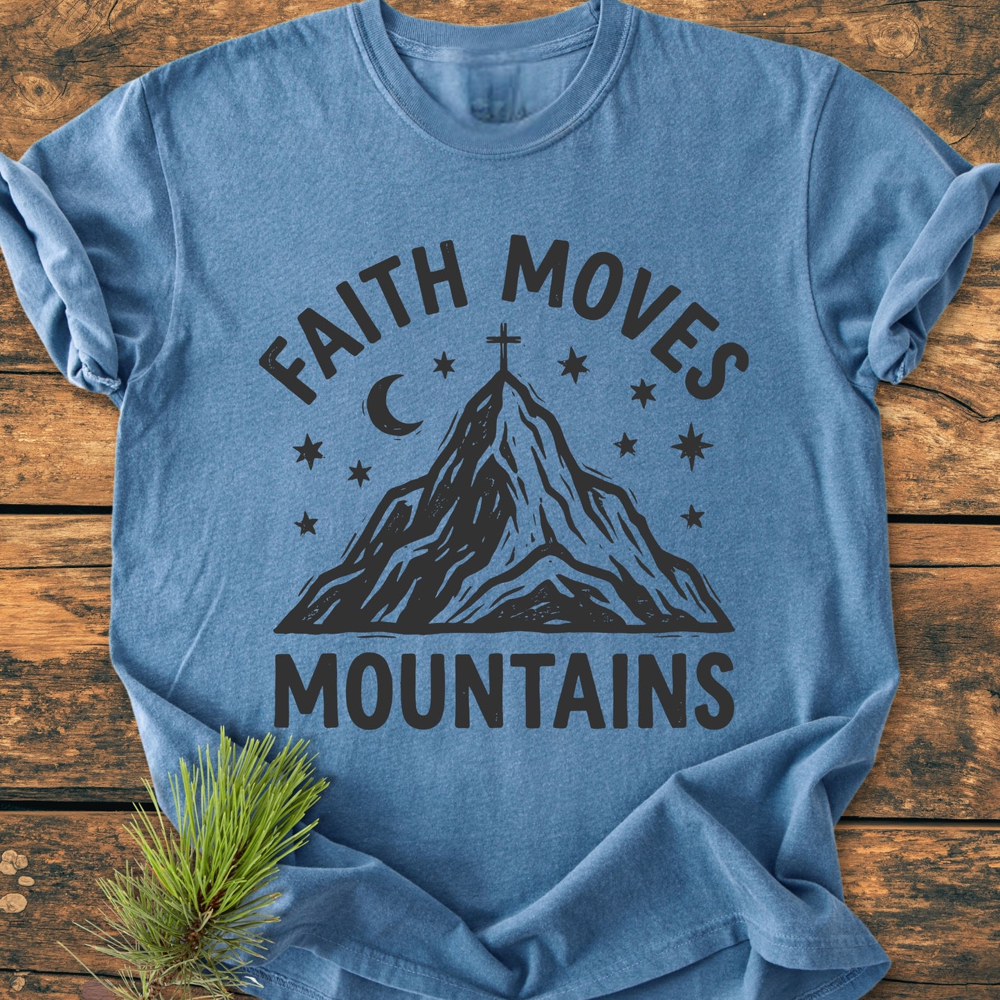 Faith Moves Mountains - Vintage Wash Tee features bold black lettering with a mountain graphic and cross. The heather-blue shirt lies on wood, accompanied by a pine sprig, showcasing its cozy, relaxed fit.
