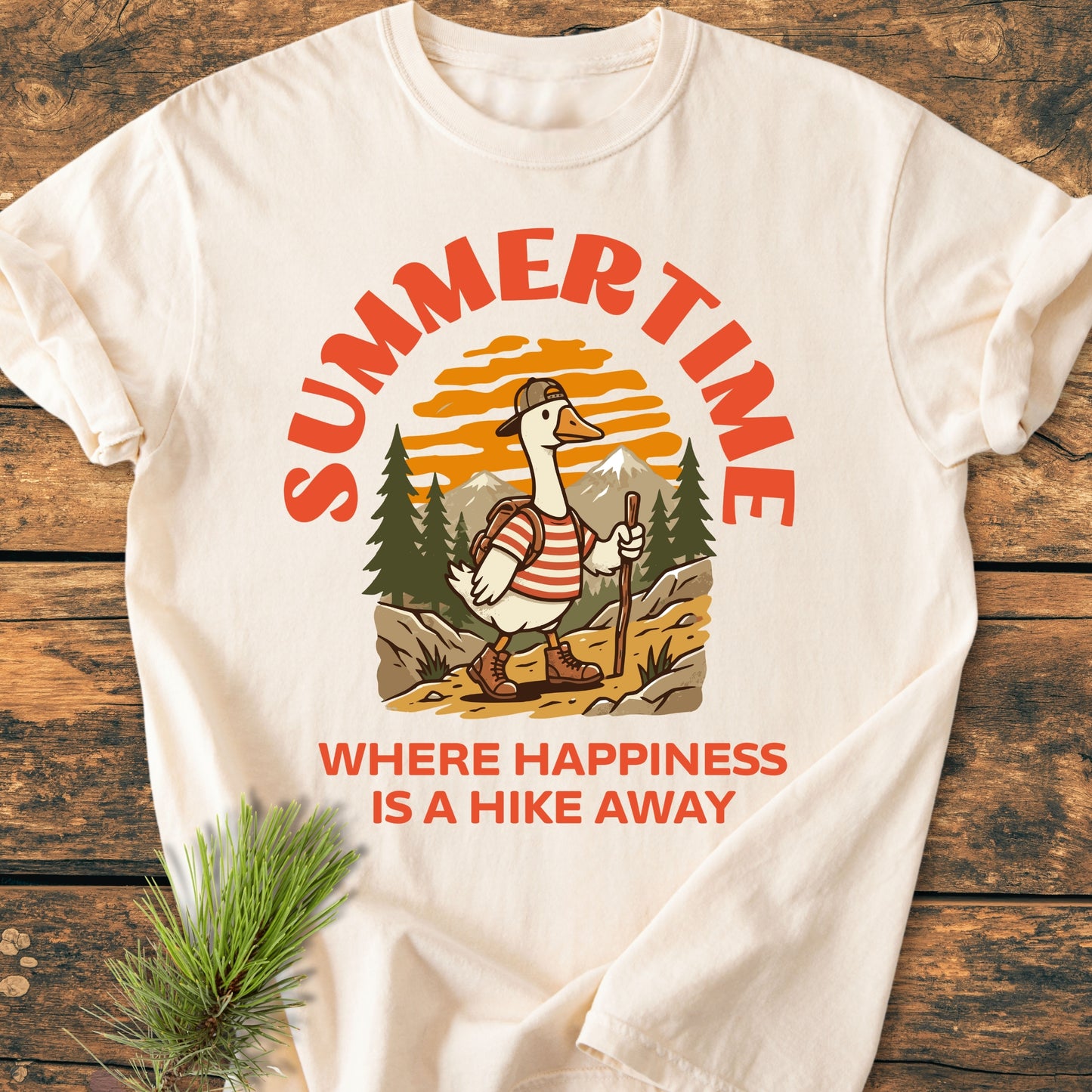 Summertime Hike - Vintage Wash Tee featuring a cartoon hiking goose with boots and backpack, against a mountain sunset, with slogans celebrating outdoor joy. Laid on a wooden surface with a pine sprig.