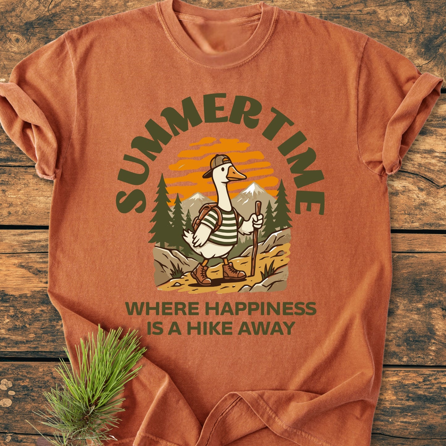 Summertime Hike - Vintage Wash Tee featuring a hiking goose graphic with a backpack on a scenic trail, laid on wood with a pine sprig beside it, showcasing its cozy, relaxed fit.