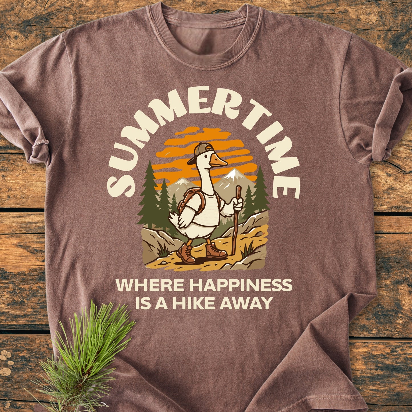 Summertime Hike - Vintage Wash Tee features a cartoon goose hiking past mountains and pine trees, with SUMMERTIME text. Laid on a wooden surface with a pine sprig accent.