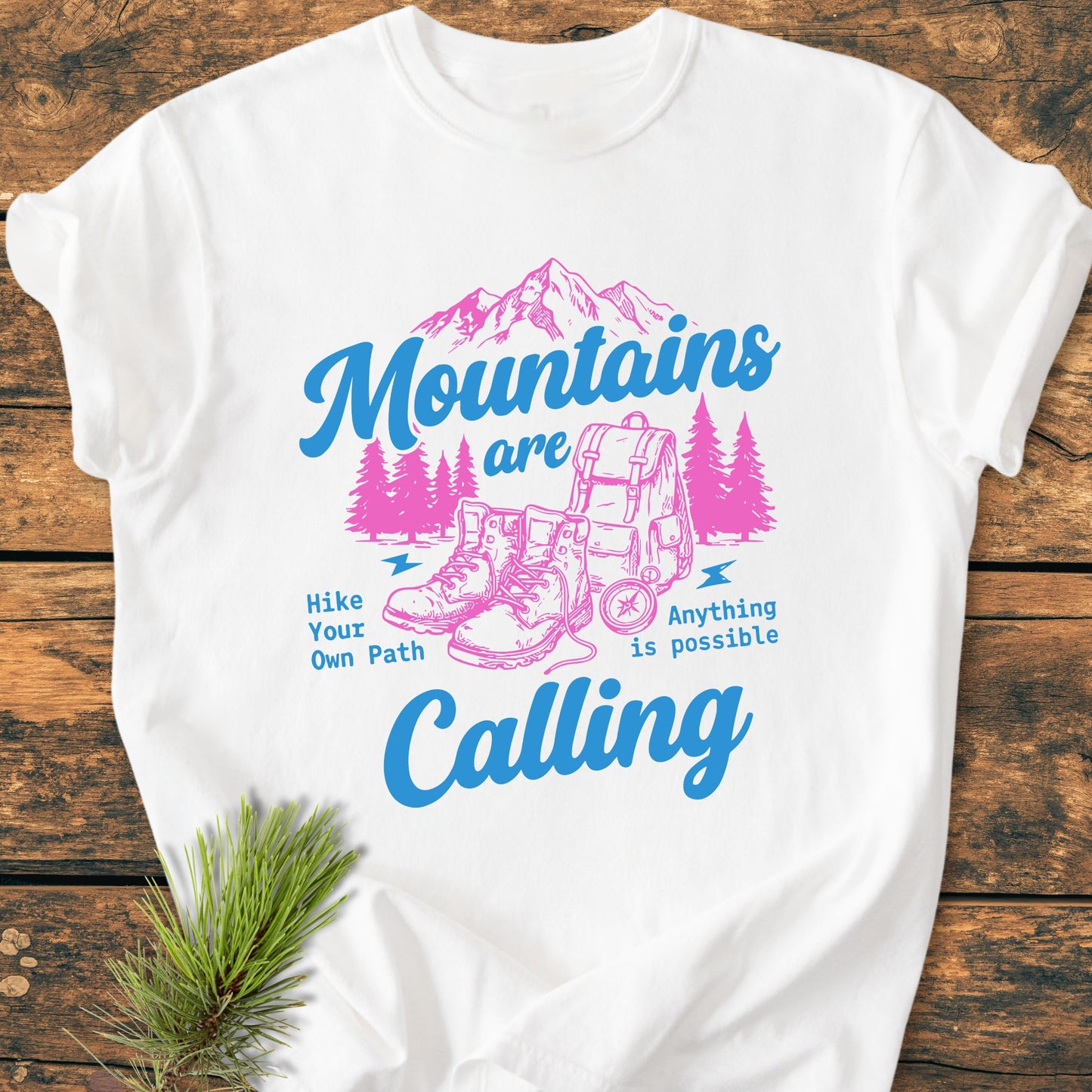 Cute Mountains Are Calling Illustration - Vintage Wash Tee features a stylized mountain range, pine trees, hiking boots, and rucksack graphic, with inspiring blue script, laid on a wooden surface.