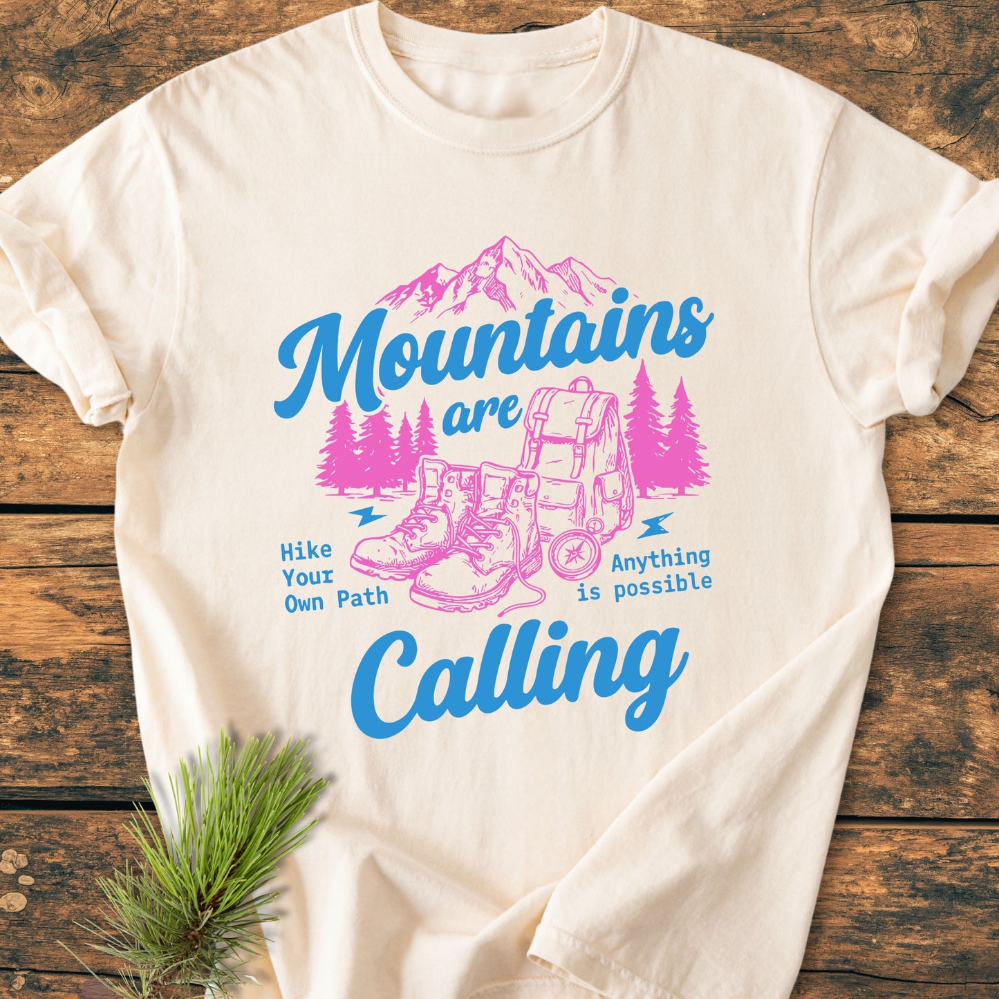 Cute Mountains Are Calling Illustration - Vintage Wash Tee featuring a pink and blue outdoor graphic with mountains, pine trees, hiking boots, and backpack, complemented by motivational blue text on a cream T-shirt.
