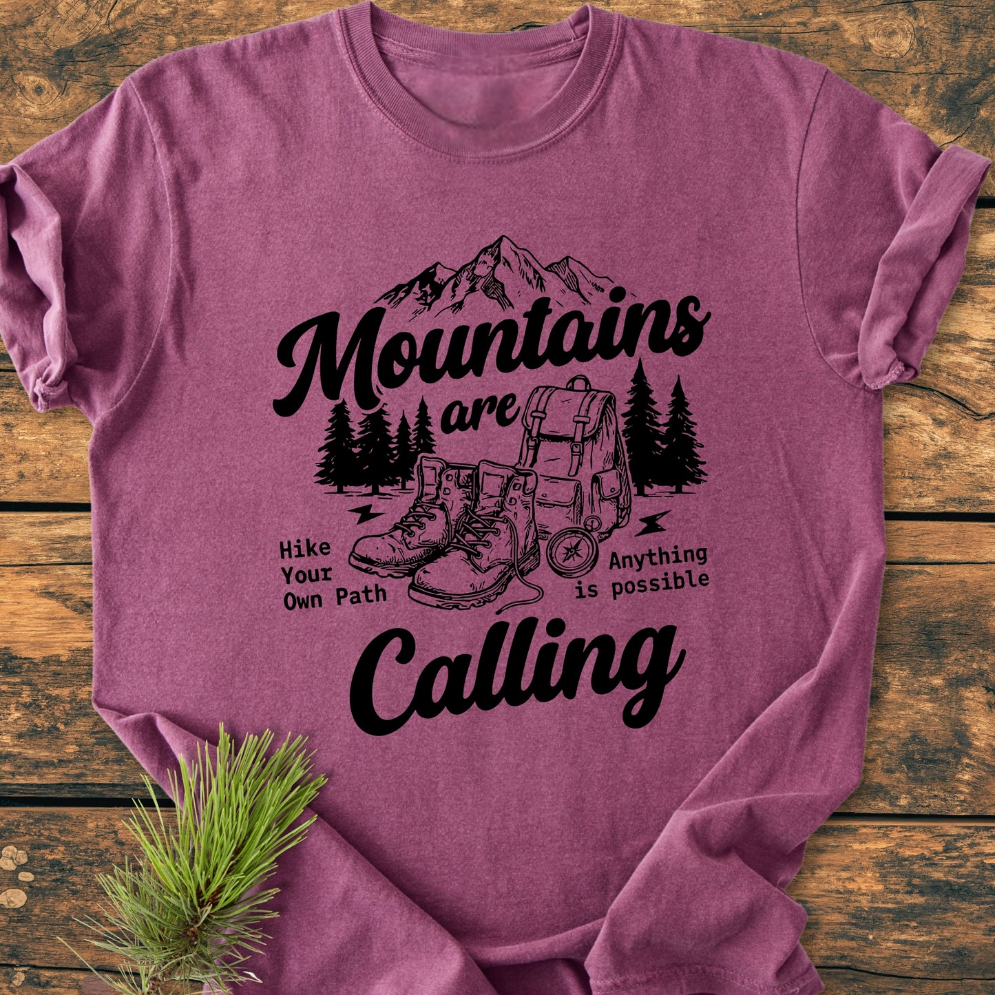 Cute Mountains Are Calling Illustration - Vintage Wash Tee: A heathered plum T-shirt with a black outdoorsy graphic of mountains, pine trees, backpack, boots, and motivational text, laid on a wooden surface.