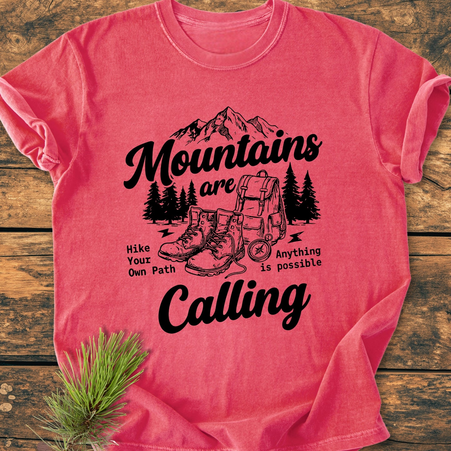 Cute Mountains Are Calling Illustration - Vintage Wash Tee features a mountain range, trees, hiking gear, and motivational slogans. Laid flat on wood with pine needles accent, showcasing its cozy, relaxed fit.