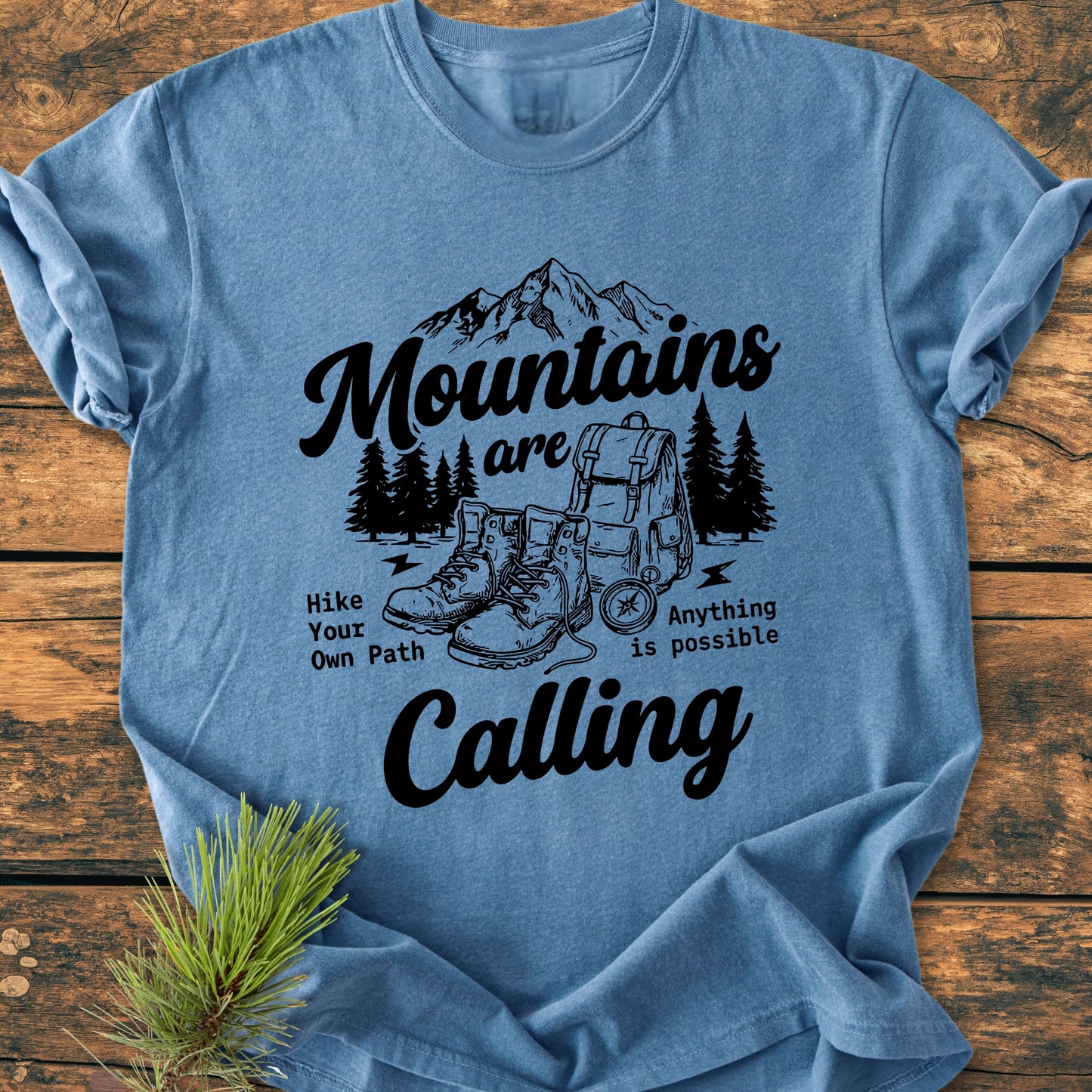 Cute Mountains Are Calling Illustration - Vintage Wash Tee featuring a mountain range, pine trees, hiking boots, and a backpack. The slogan reads “Mountains are Calling” with “Hike Your Own Path” beneath.