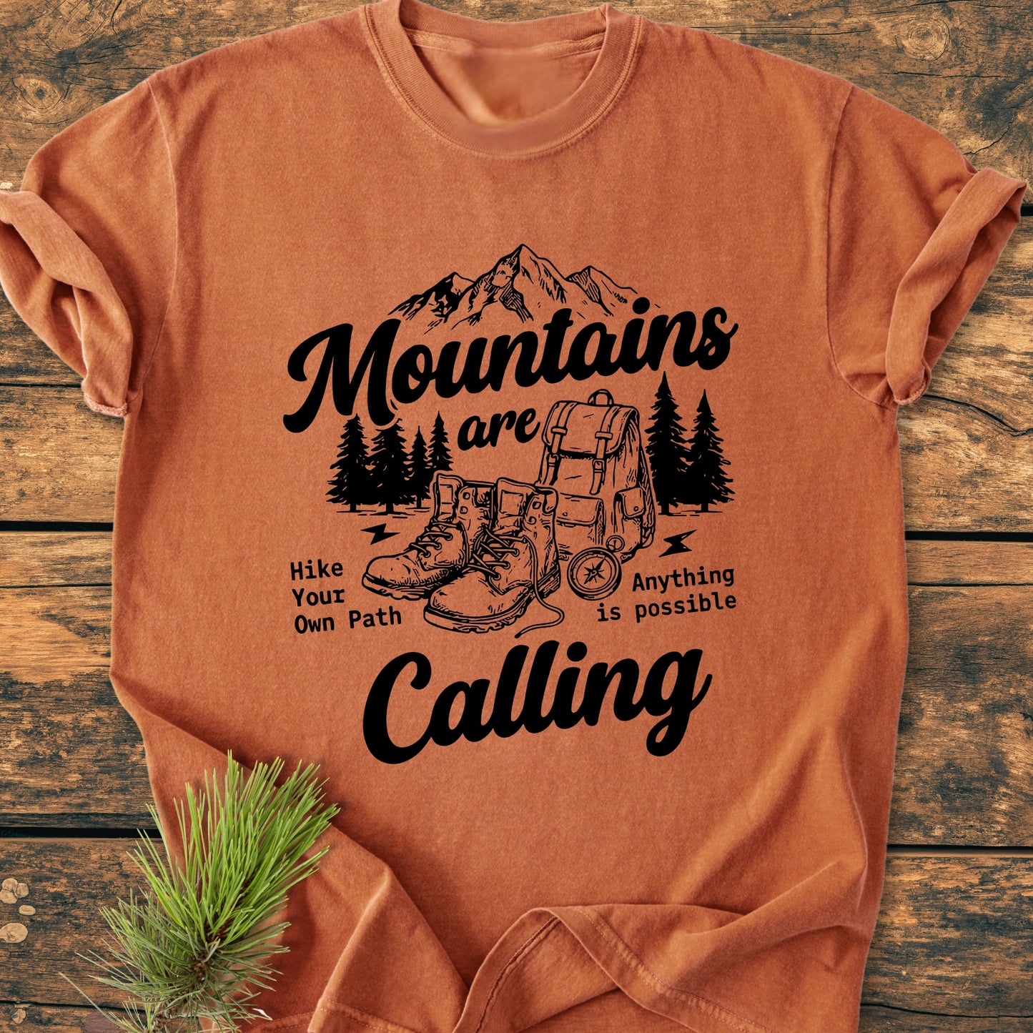 Cute Mountains Are Calling Illustration - Vintage Wash Tee features a stylized outdoor scene with mountains, pine trees, and hiking gear, accompanied by motivating taglines. Laid on wood, highlighting its relaxed, durable design.