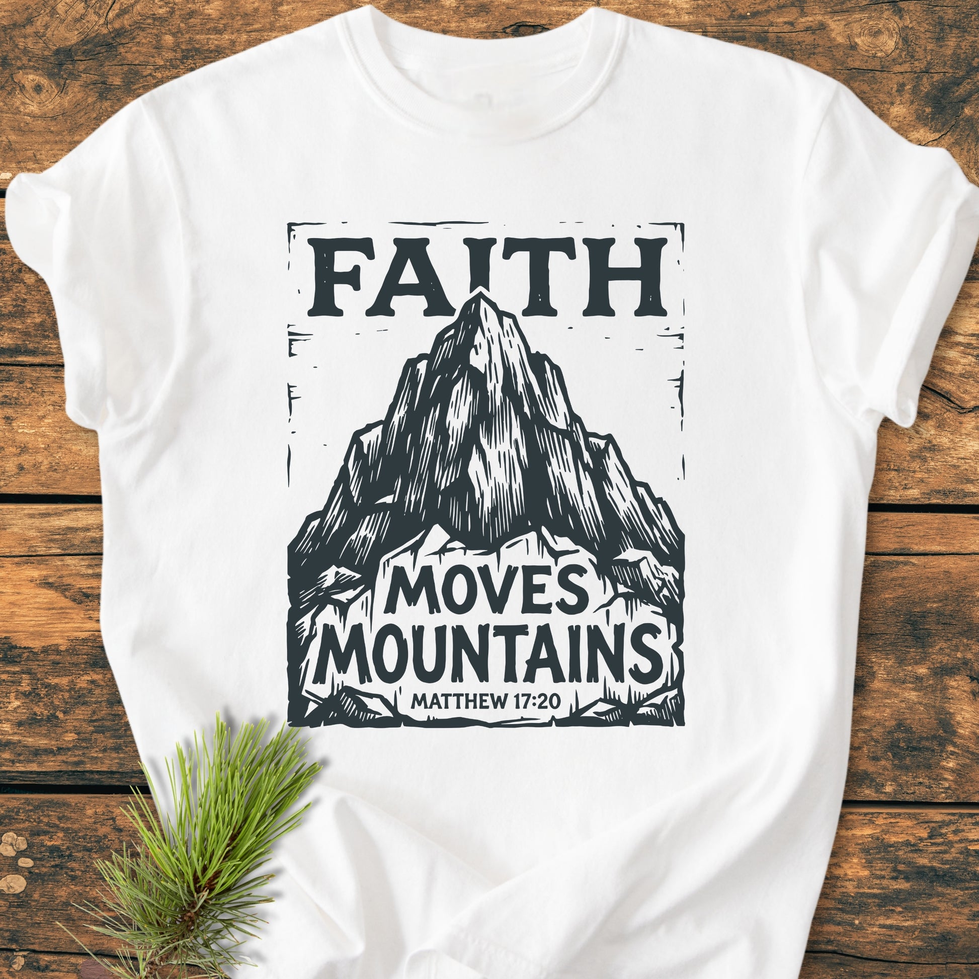 Faith Moves Mountains - Vintage Wash Tee features a mountain peak graphic on a white T-shirt, capturing an adventurous spirit. Laid on wood with pine sprig, it embodies rustic charm and inspirational faith.