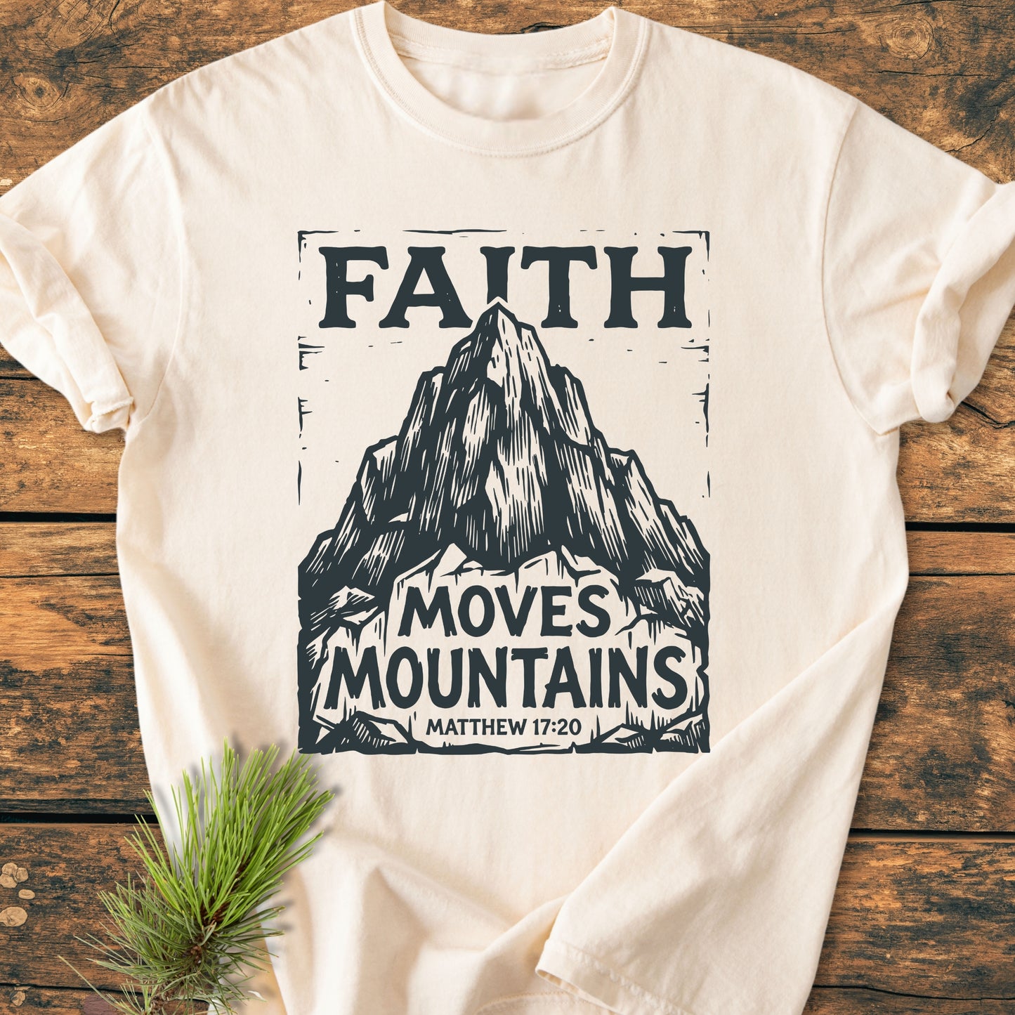 Faith Moves Mountains - Vintage Wash Tee featuring a stylized mountain graphic and biblical text, laid on wood with pine sprig; cozy, relaxed fit from 100% ring-spun cotton, emphasizing durability and comfort.