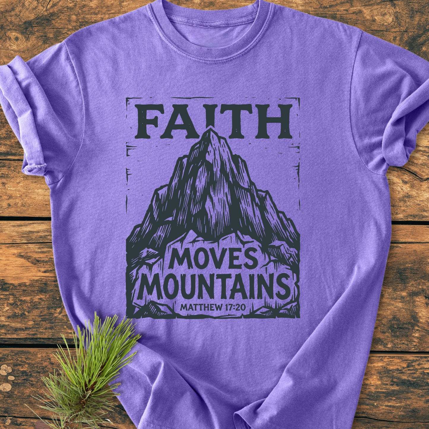 Faith Moves Mountains - Vintage Wash Tee featuring a black mountain design with FAITH MOVES MOUNTAINS and Matthew 17:20, displayed on a wooden surface with a pine sprig nearby.