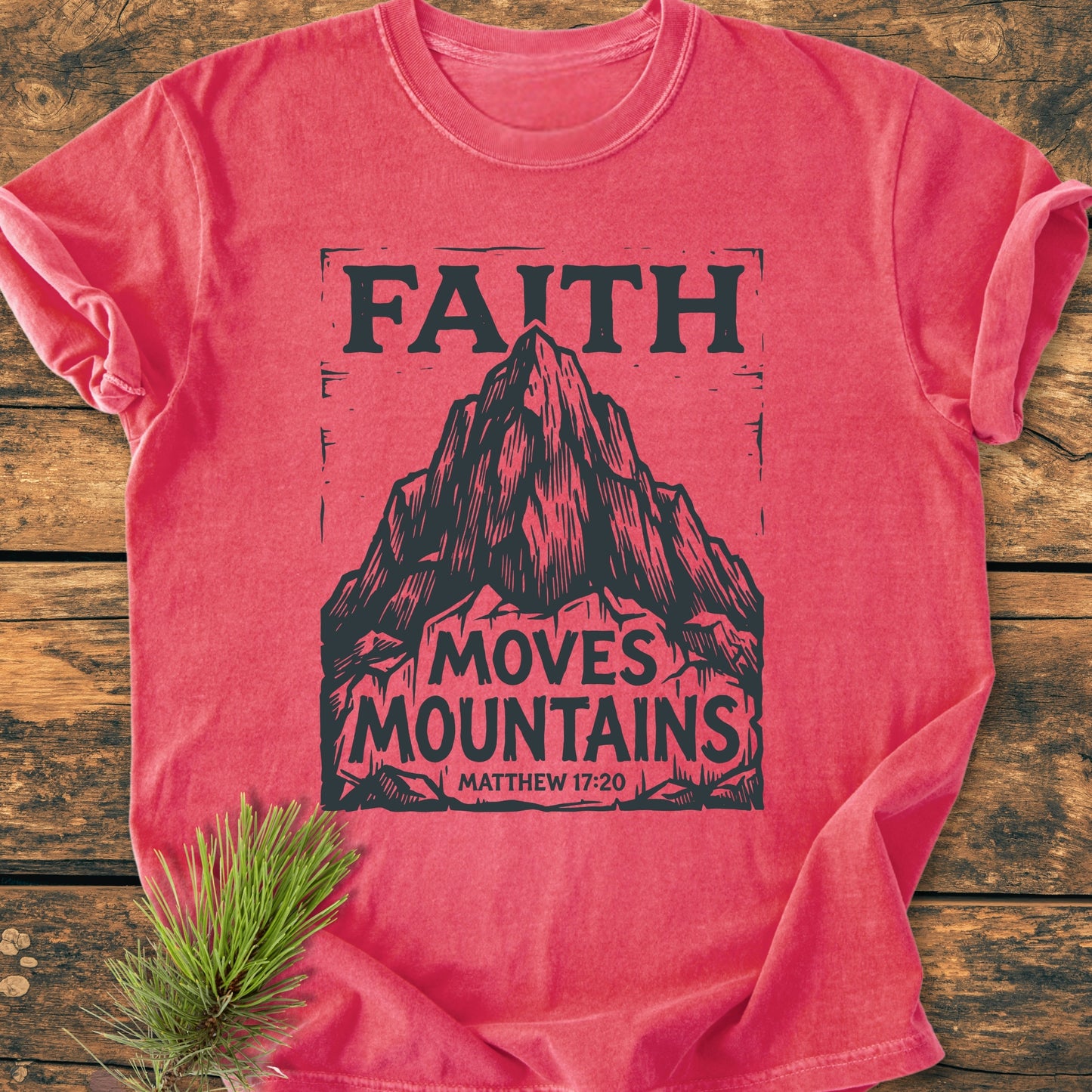 Faith Moves Mountains - Vintage Wash Tee features a stylized mountain graphic and “Matthew 17:20” on a heather-red T-shirt, laid on wood, with a pine sprig accenting its cozy, durable design.