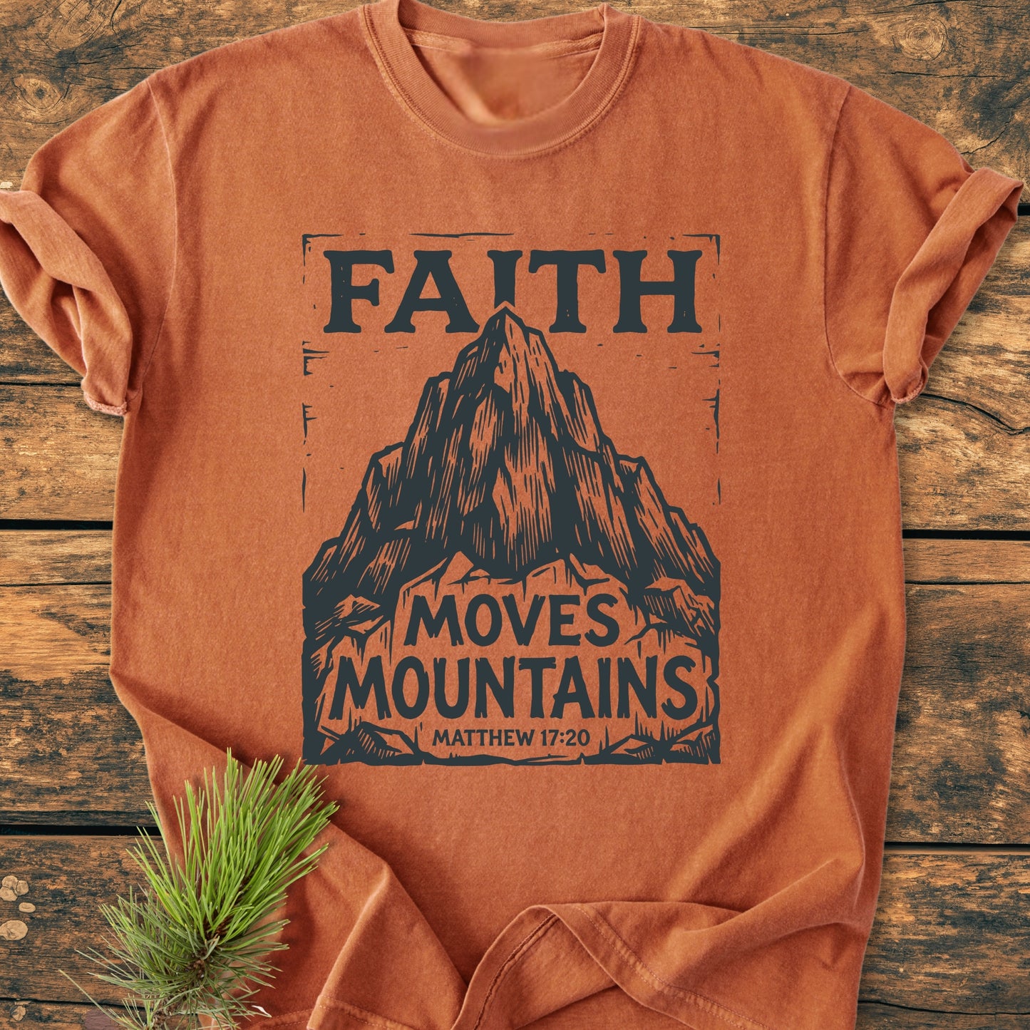 Faith Moves Mountains - Vintage Wash Tee in rust-orange, featuring a black mountain graphic with FAITH MOVES MOUNTAINS and Matthew 17:20, displayed on a wooden surface with a pine sprig beside it.