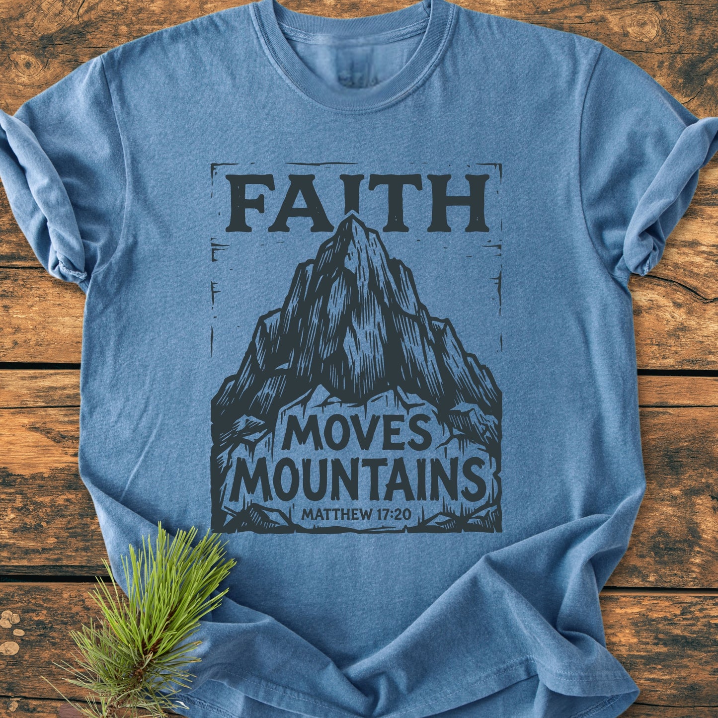 Faith Moves Mountains - Vintage Wash Tee displayed on rustic wood, features a mountain graphic with biblical text. Heather-blue, crew-neck, known for softness and relaxed fit, complemented by a pine sprig.
