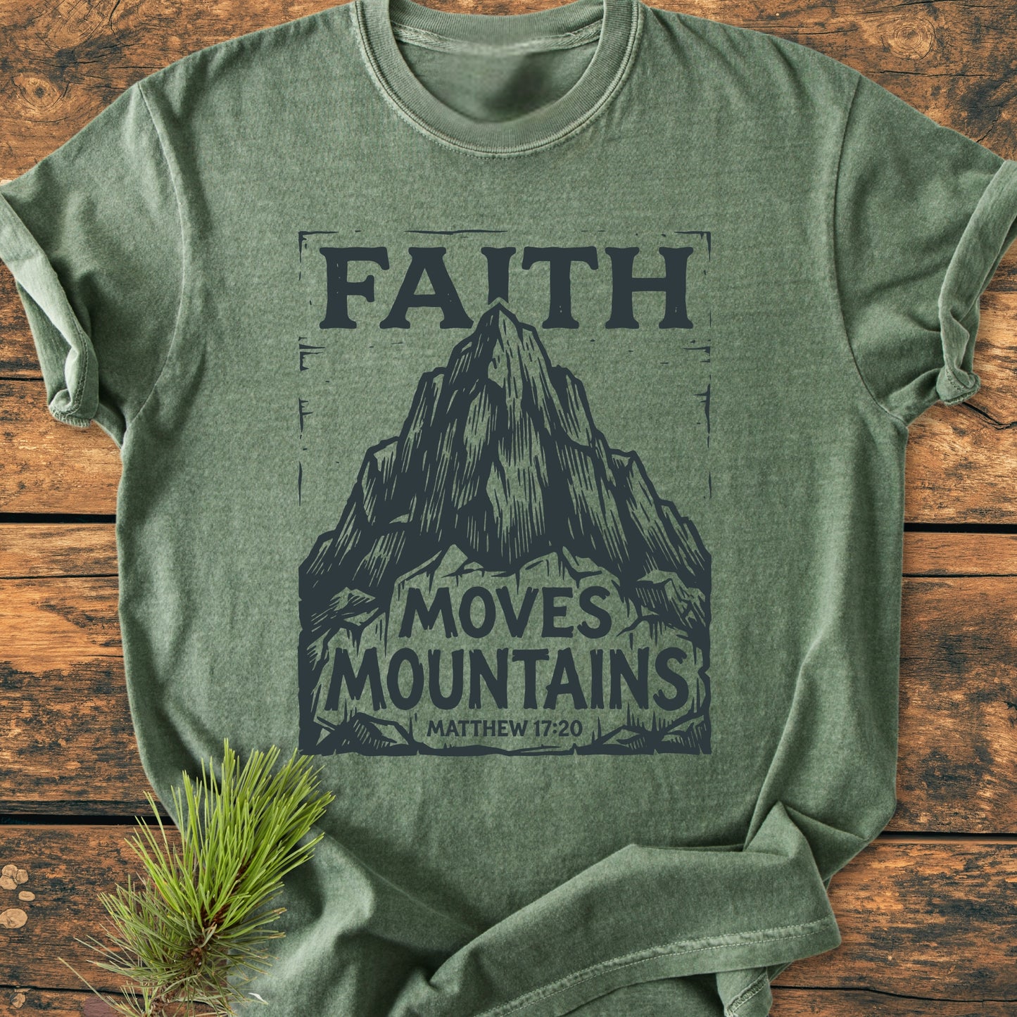 Faith Moves Mountains - Vintage Wash Tee: A green heathered T-shirt with a mountain graphic and FAITH MOVES MOUNTAINS text, featuring Matthew 17:20, laid flat with a pine branch accent.