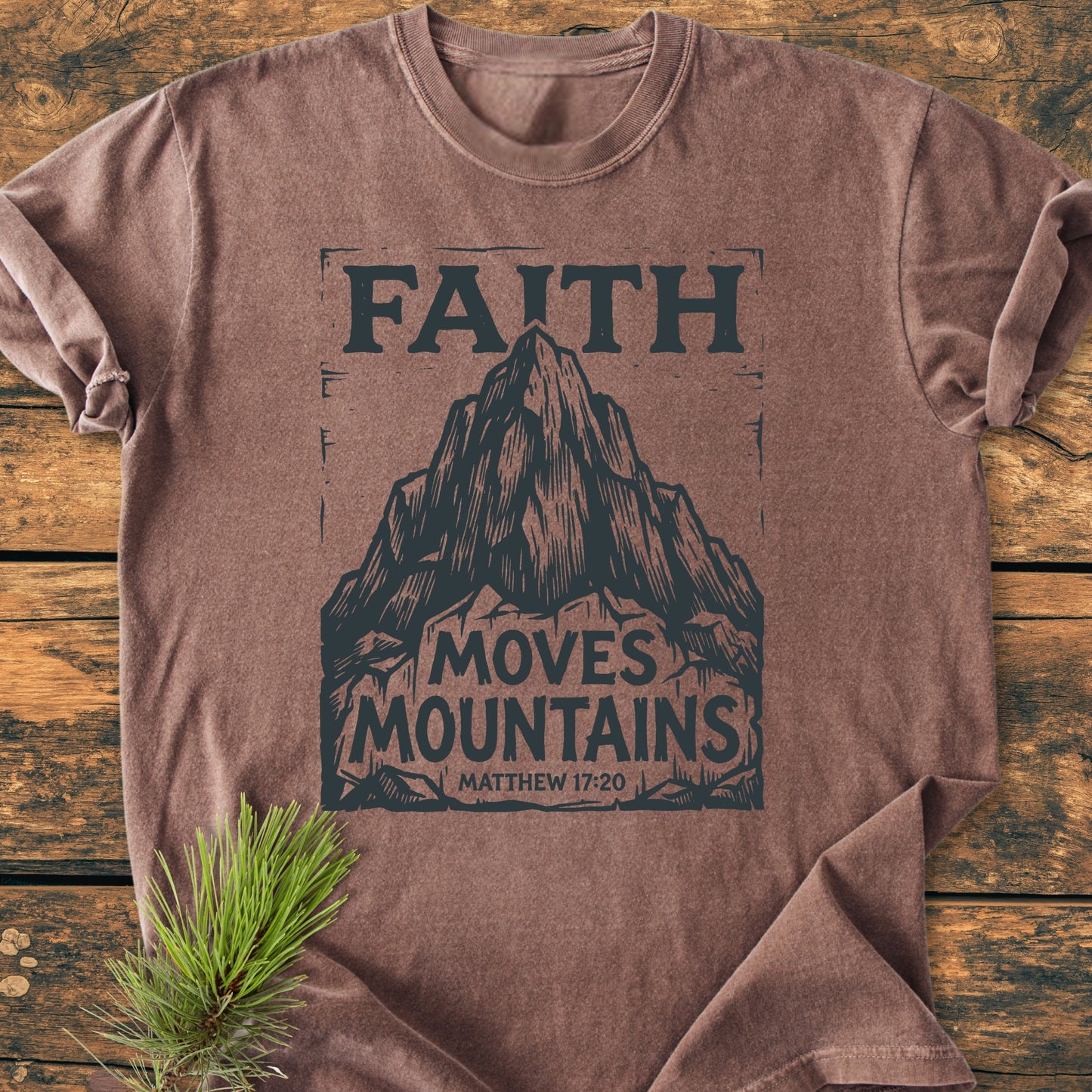 Faith Moves Mountains - Vintage Wash Tee featuring a mountain graphic and scripture reference. Laid on wood with a pine sprig, showcasing its relaxed fit and garment-dyed softness.