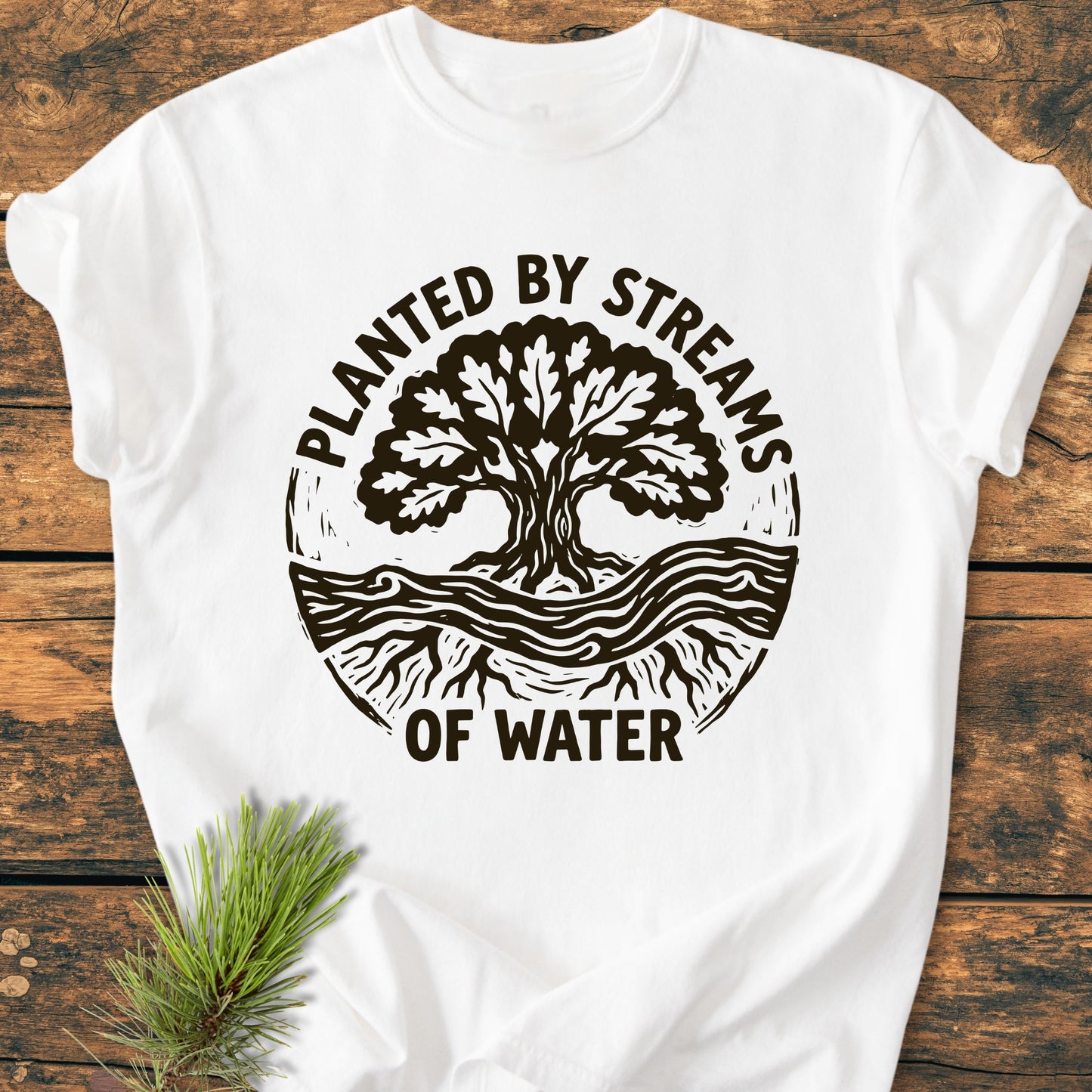 Planted By Streams, Psalm 1,3 - Vintage Wash Tee features a bold circular graphic with a tree, stream, and roots, laid on wood with pine needles, highlighting its soft, durable, relaxed fit.