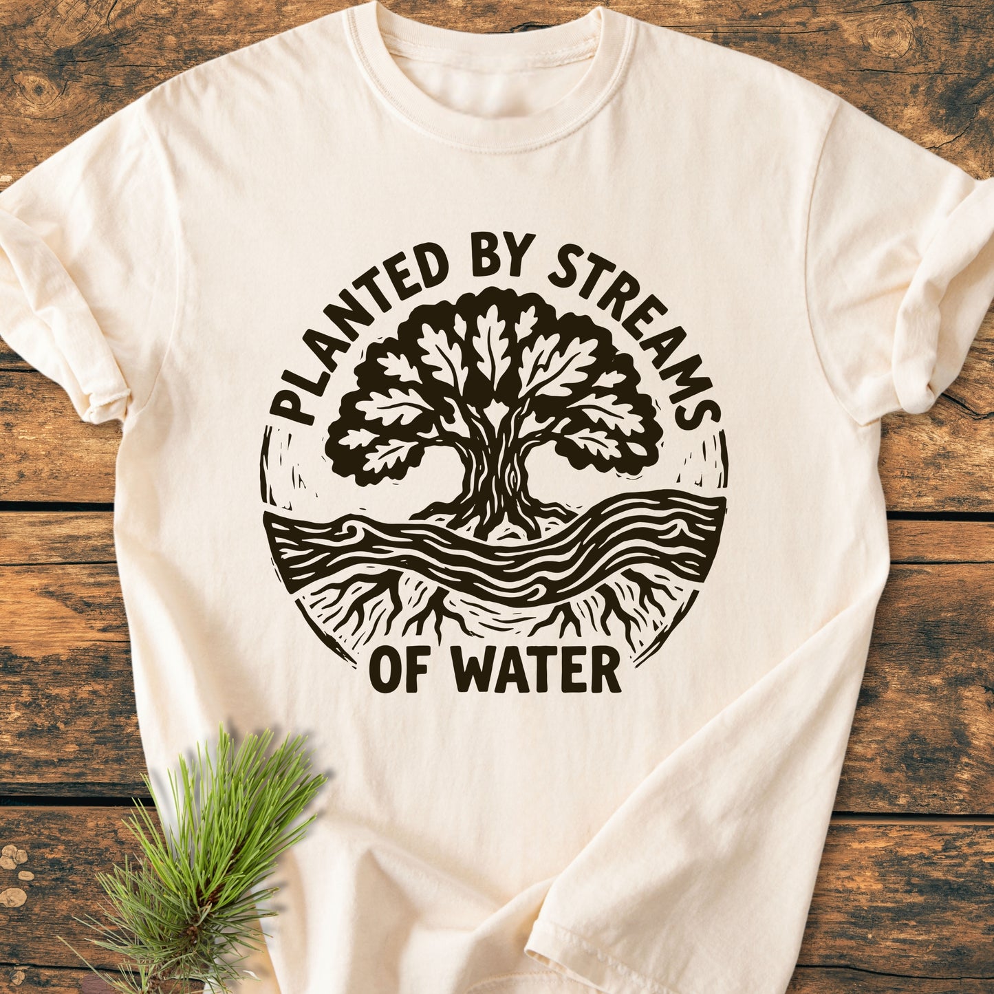 Planted By Streams, Psalm 1,3 - Vintage Wash Tee features a tree and stream illustration on a cream T-shirt, laid on wood with a pine sprig, highlighting its rustic and natural appeal.
