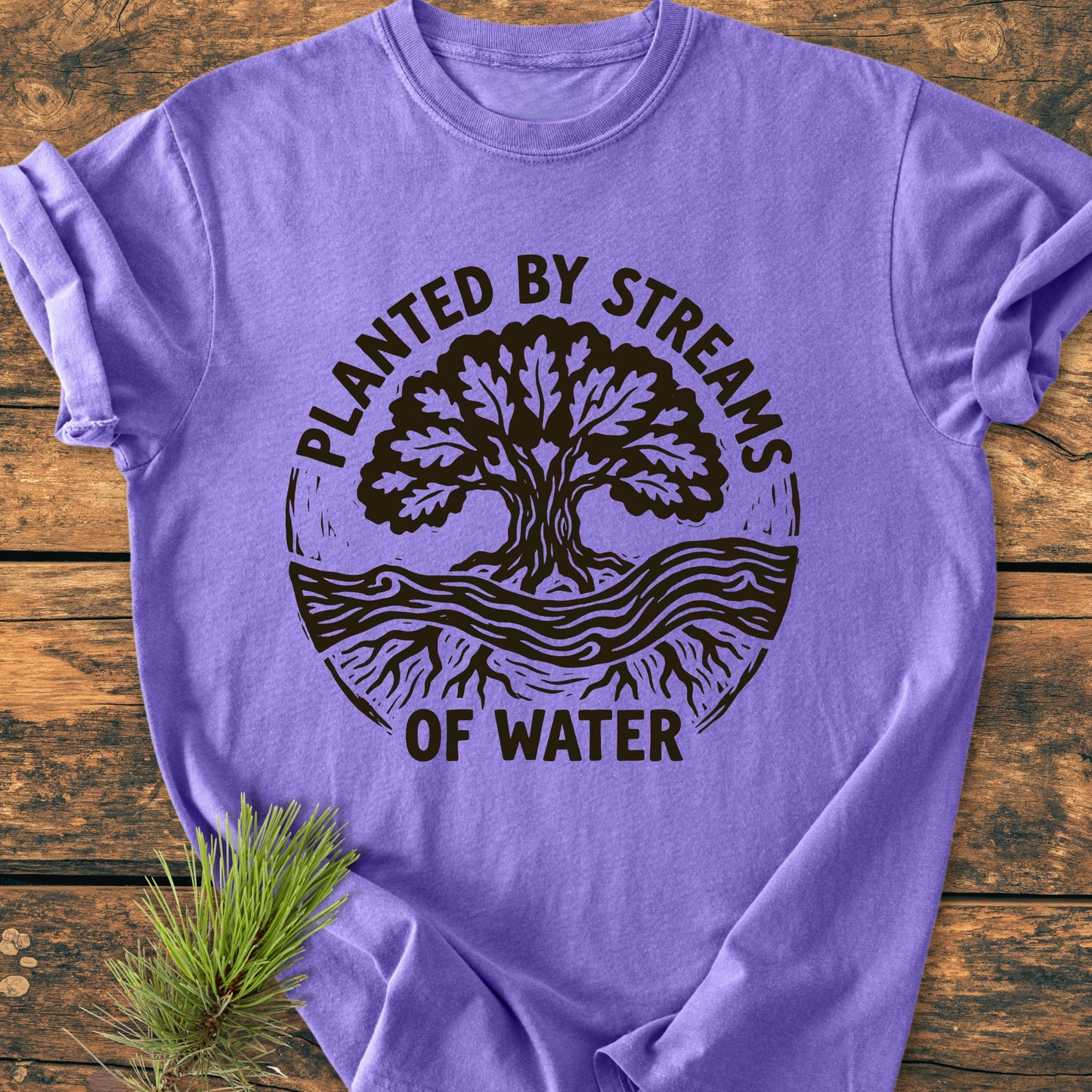 Planted By Streams, Psalm 1,3 - Vintage Wash Tee features a circular tree graphic with roots and a stream on a lavender-purple T-shirt, laid on a wooden tabletop with a pine sprig.